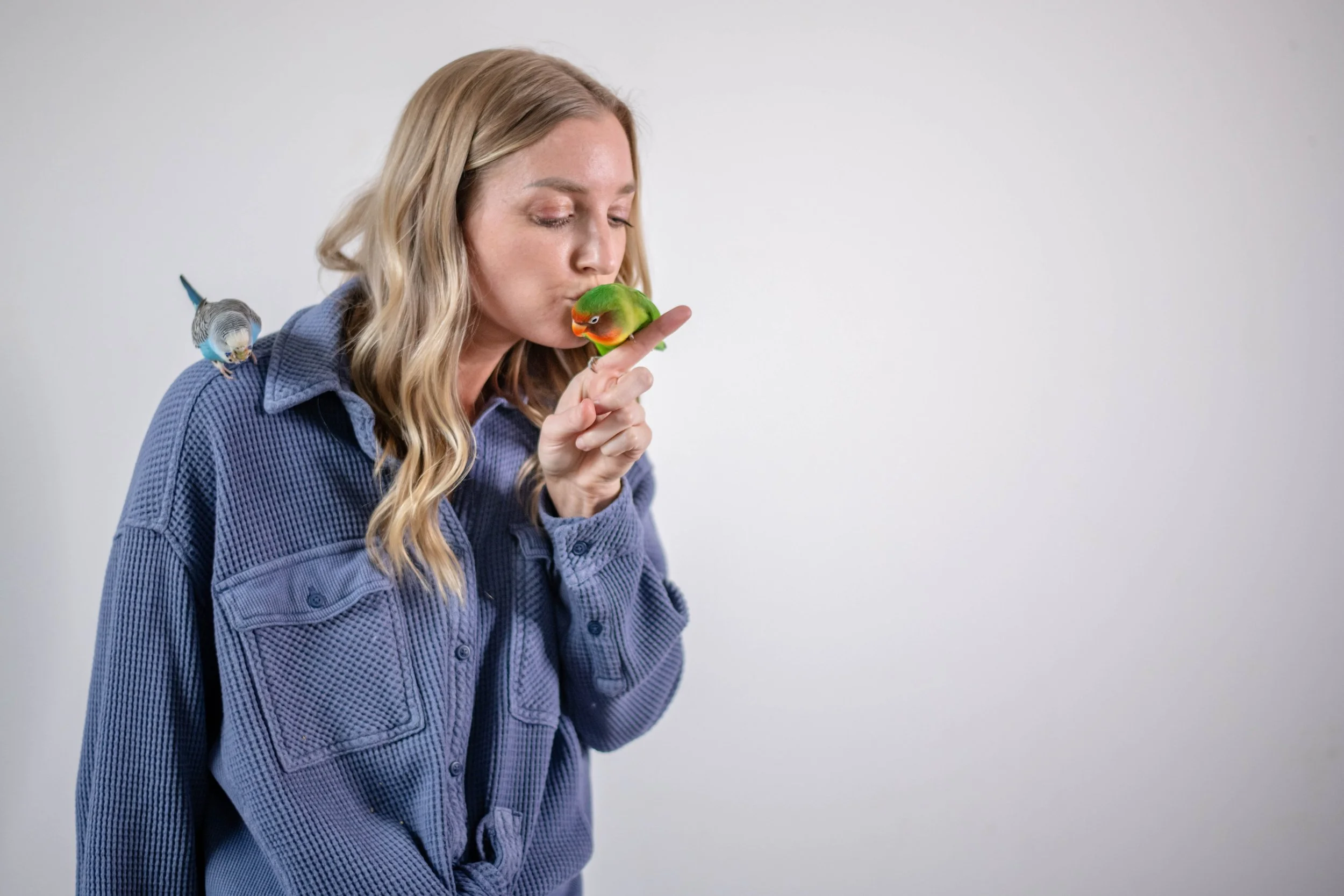 Woman in blue plaid shirt kissing a small green and orange bird on her finger, with another small bird perched on her shoulder, against a plain white background.
