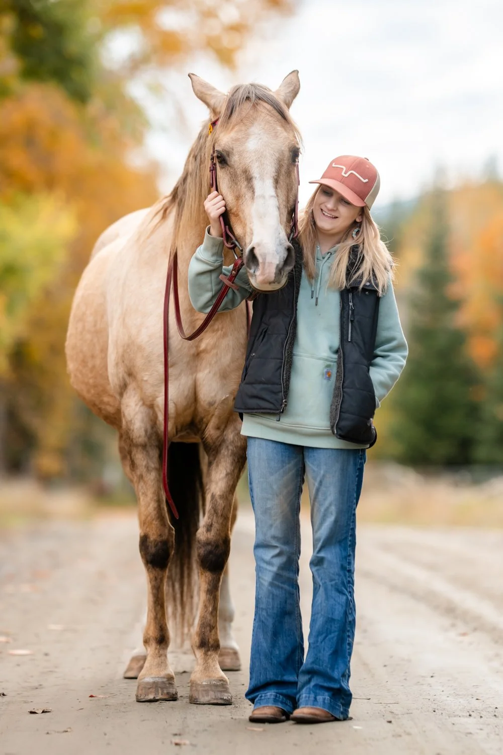 A young woman with blonde hair, wearing a pink cap, gray hoodie, and black vest, stands next to a light brown horse with a white stripe on its face, on a dirt path lined with trees with autumn-colored leaves.