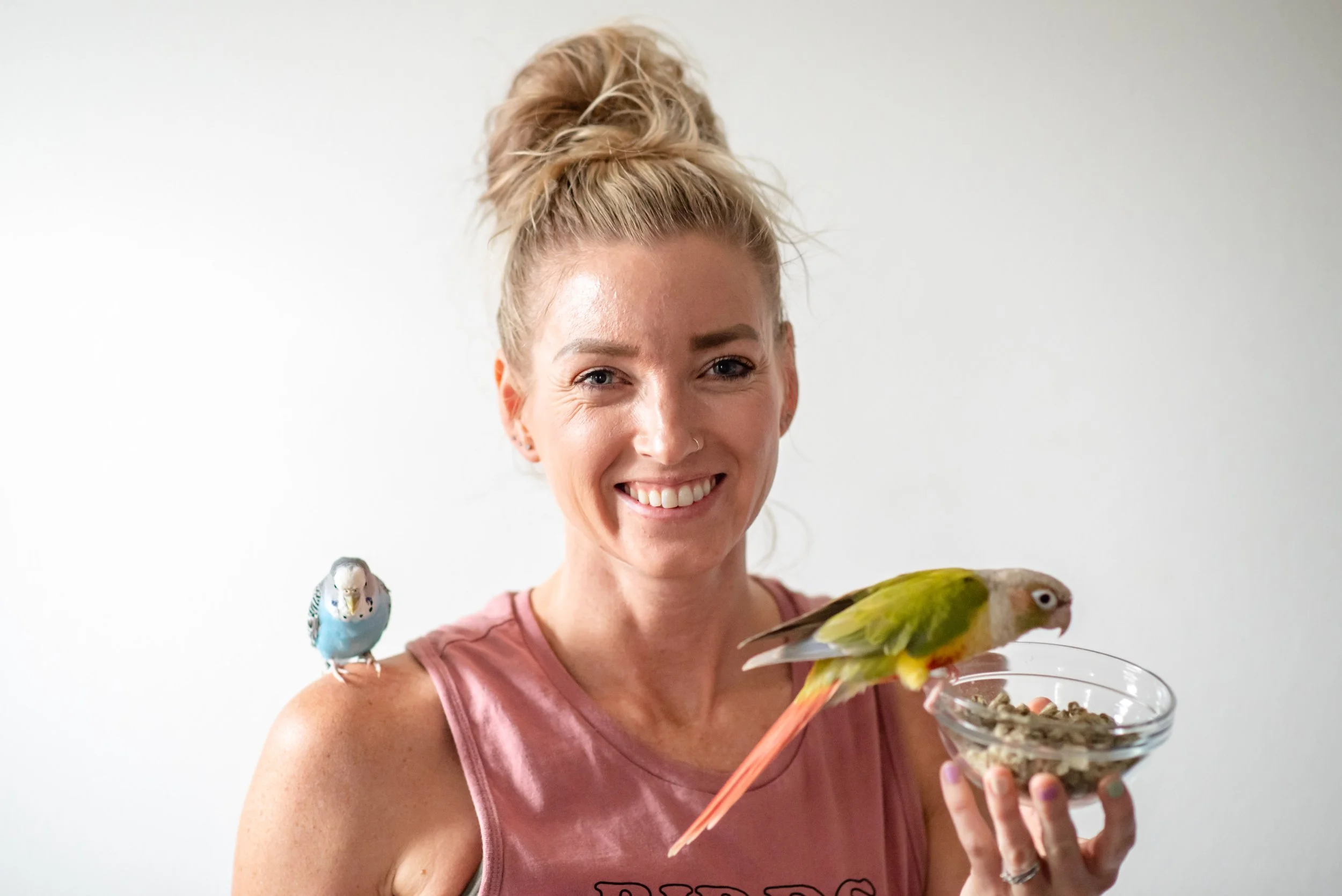 A smiling woman with blonde hair in a messy bun, wearing a pink tank top, holding a glass bowl of birdseed with a green and yellow parrot perched on her finger and a blue budgerigar sitting on her shoulder.