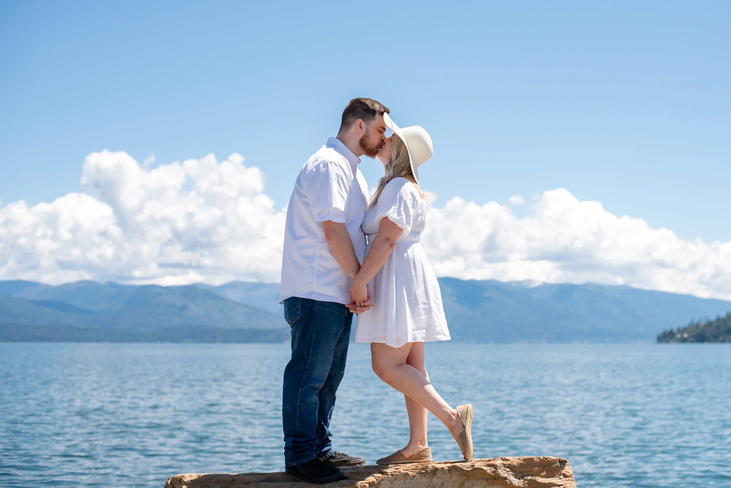 A couple kissing on a log by Lake Pend Oreille, with mountains and a blue sky with white clouds in the background.