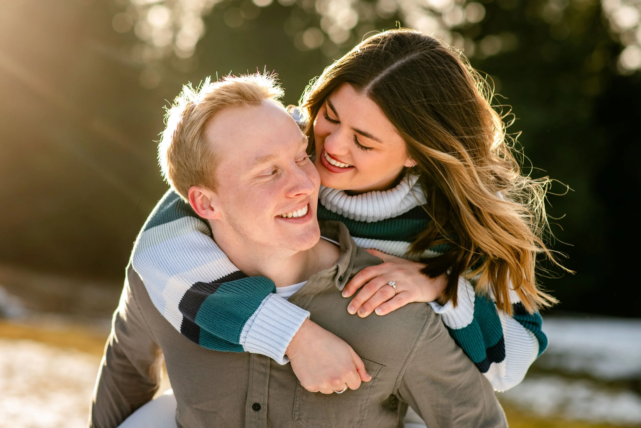 A happy couple outdoors enjoying a piggyback ride near a body of water during golden hour, smiling and looking at each other.