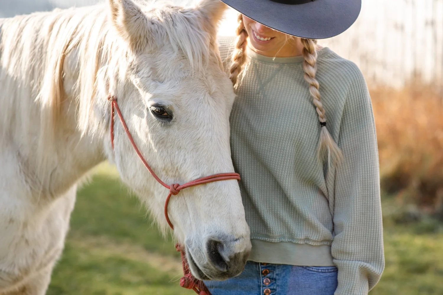 A woman with braided blonde hair wearing a wide-brimmed hat and a light green sweater is standing next to a white horse with a red halter, both enjoying a moment outdoors.