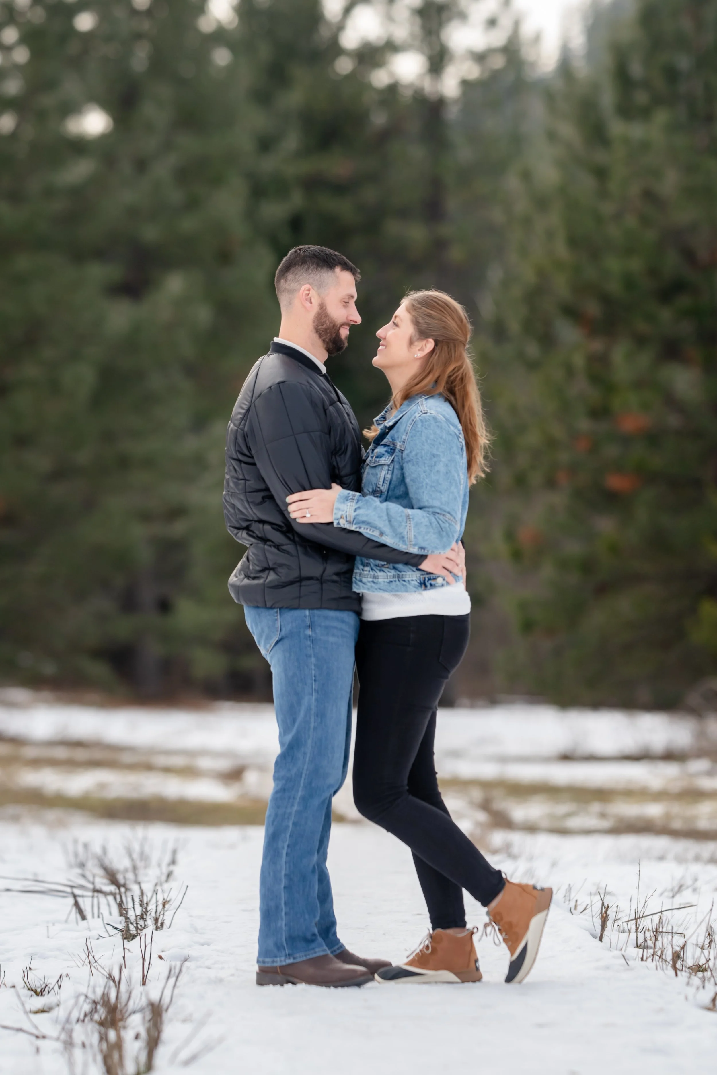 A couple standing close together outdoors in a snowy area, embracing and looking into each other's eyes with trees in the background.