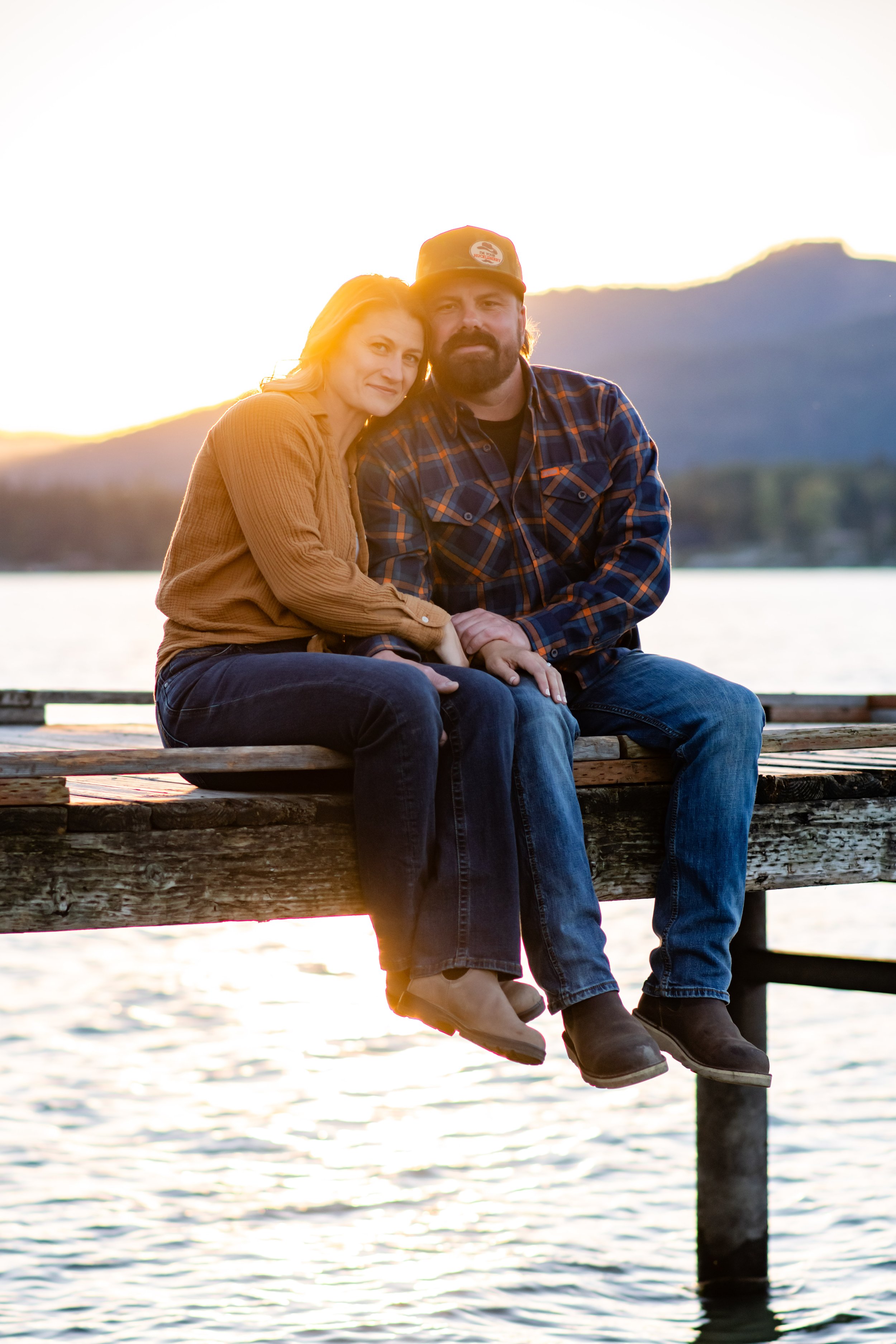 A couple sitting on a wooden dock at sunset near Lake Pend Oreille with mountains in the background, holding hands and smiling.