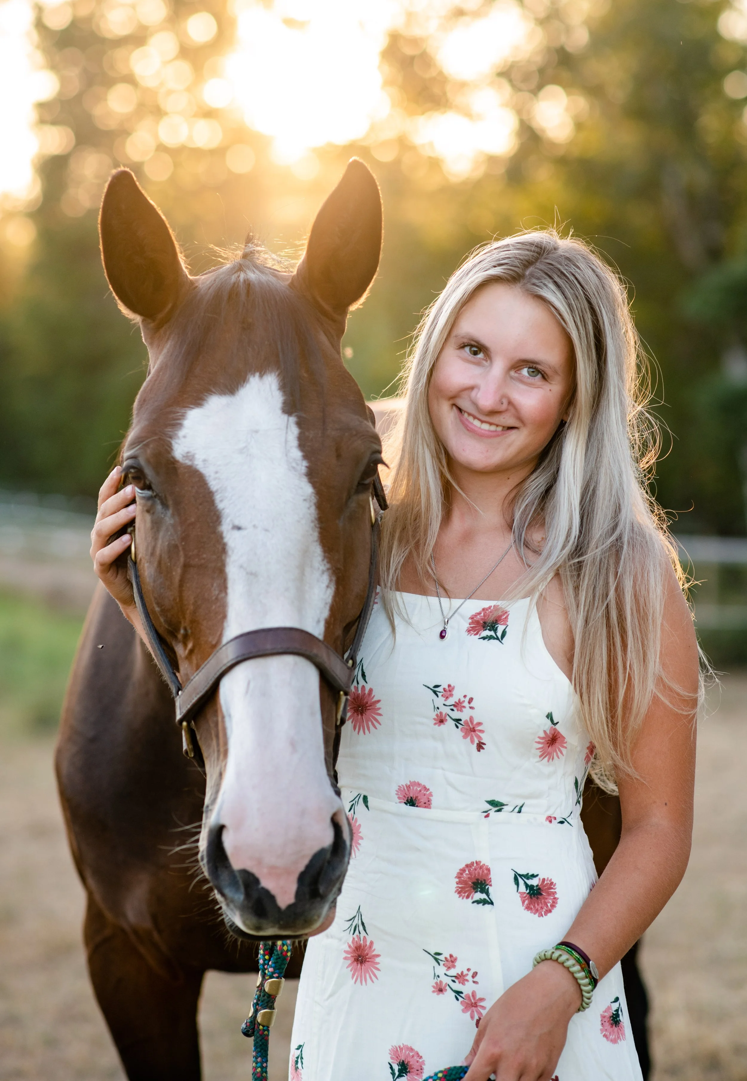A smiling young woman with long blonde hair standing beside a horse, holding its bridle, outdoors during sunset.