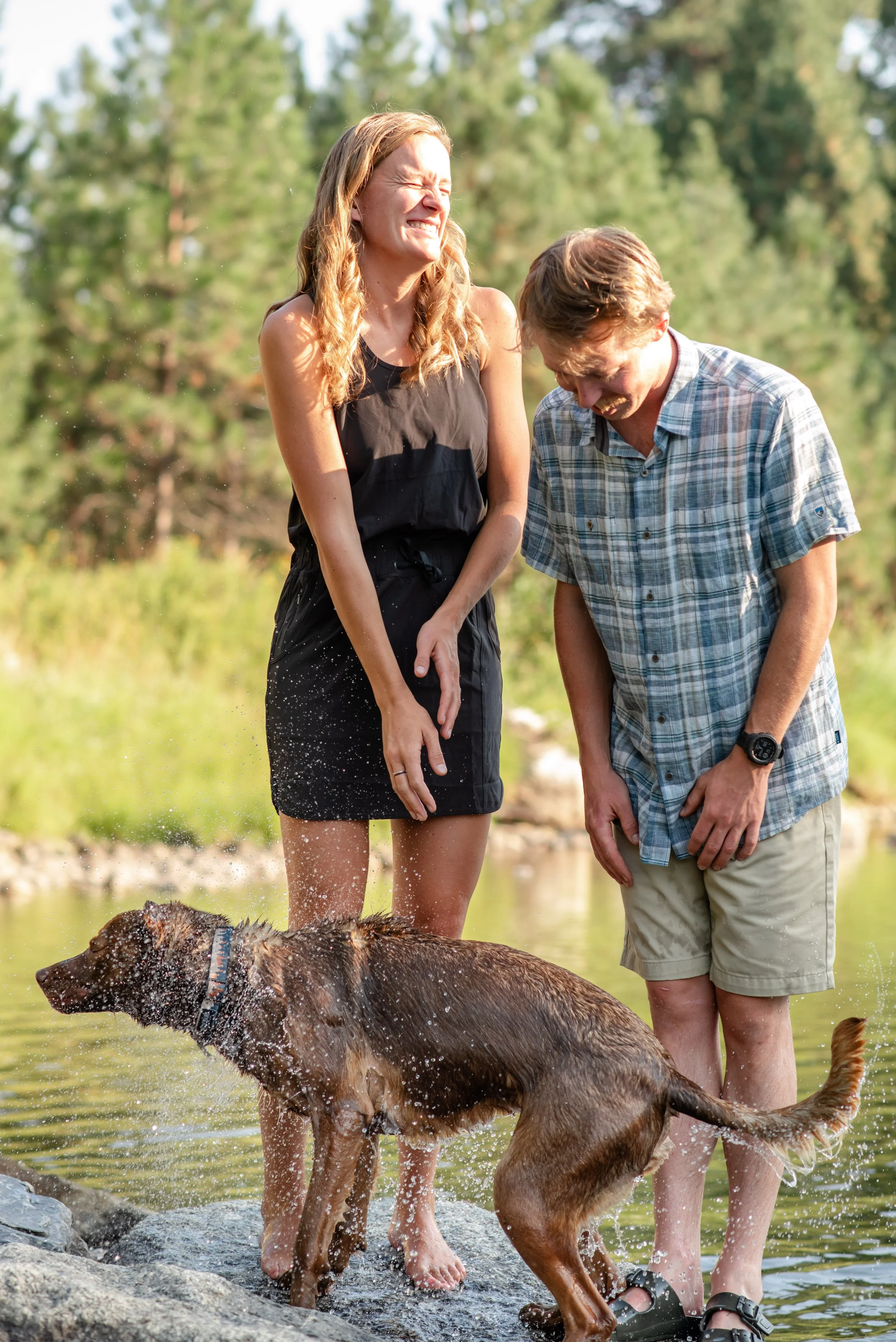 A woman and a man laughing and wet from a stream while a dog plays in the water