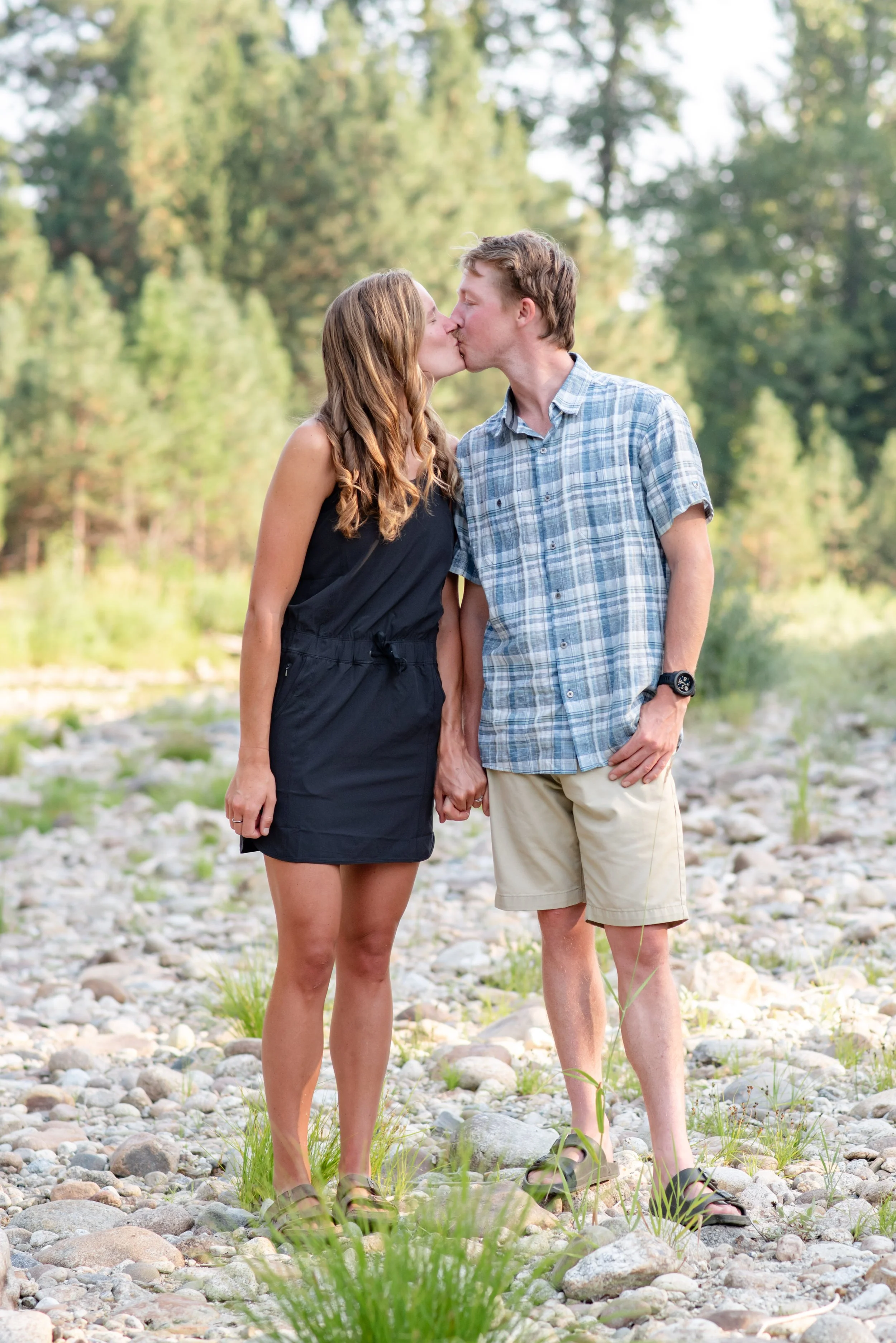 A couple kissing outdoors on a rocky area with trees in the background, holding hands.