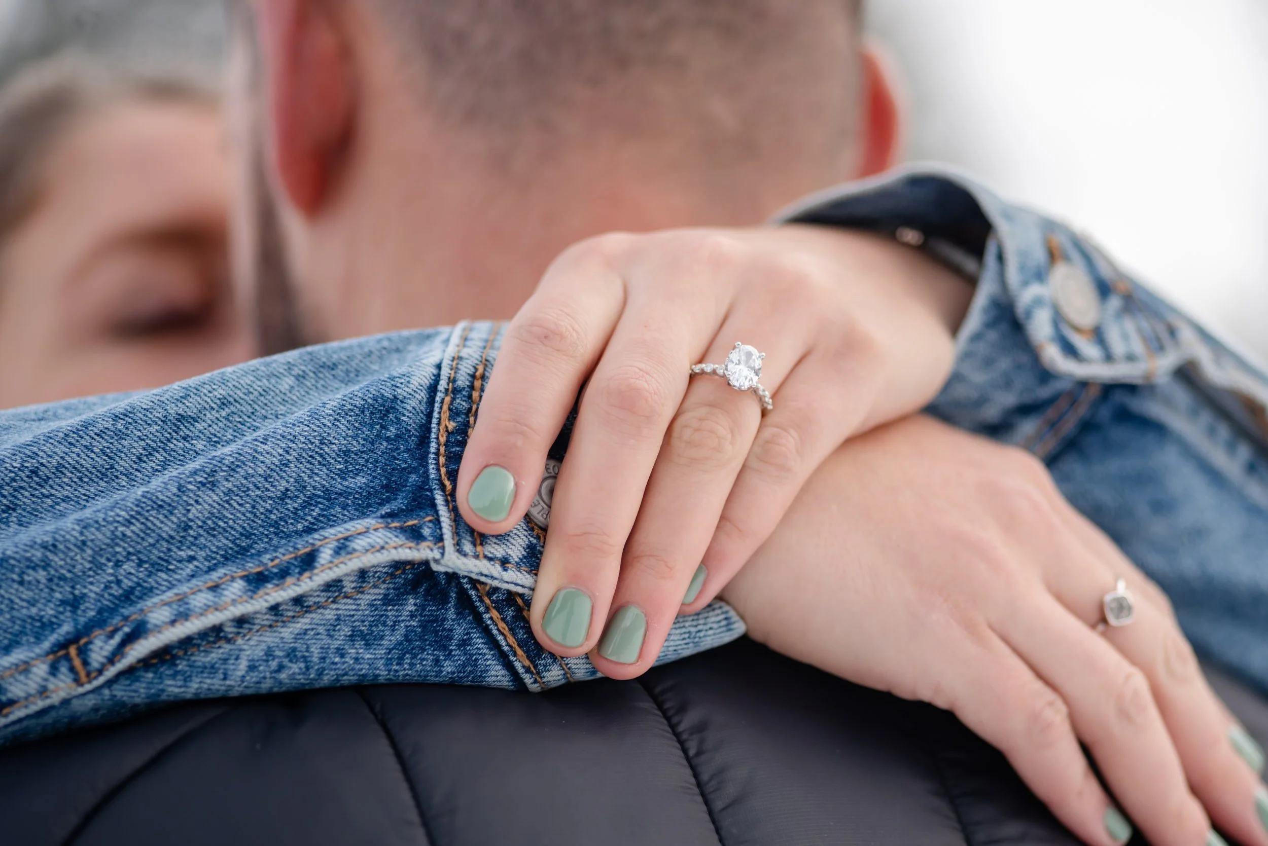 A woman with mint green nail polish wearing an engagement ring on her finger, resting her hand on a man's shoulder. The man is wearing a denim jacket and the focus is on the rings and hands.