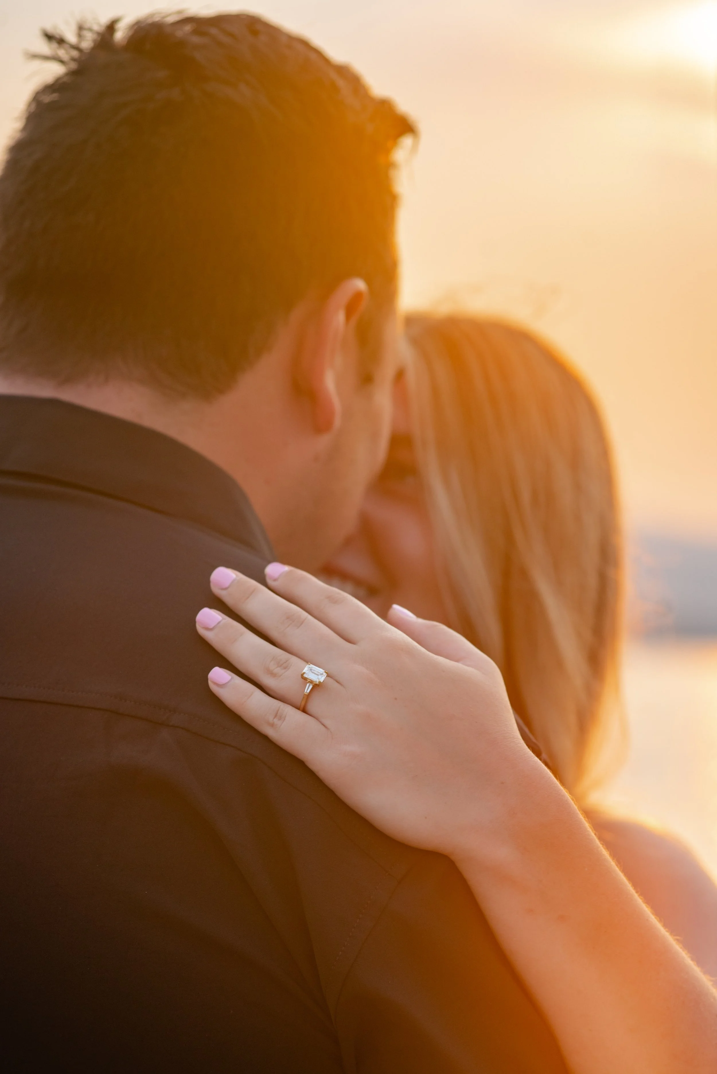 A couple embracing outdoors during sunset, with the woman showing her engagement ring on her finger.