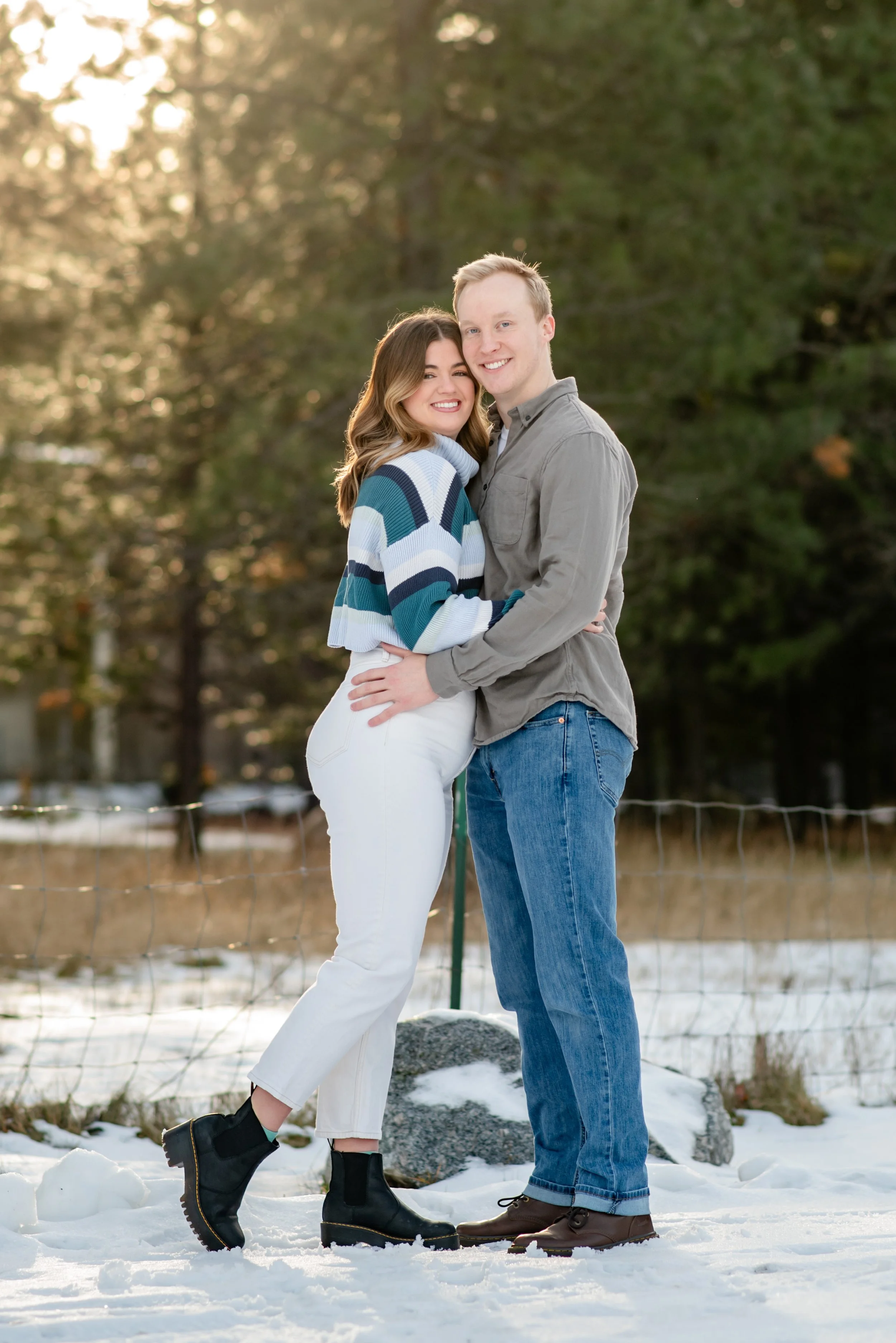 A smiling couple embracing outdoors in a snowy landscape during sunset, with trees in the background.