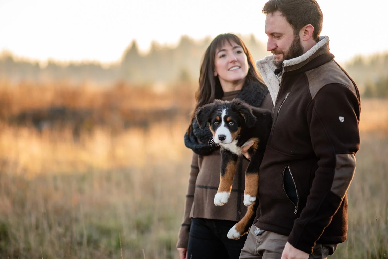 A couple outdoors holding a black, white, and tan Australian Shepherd puppy at sunset.