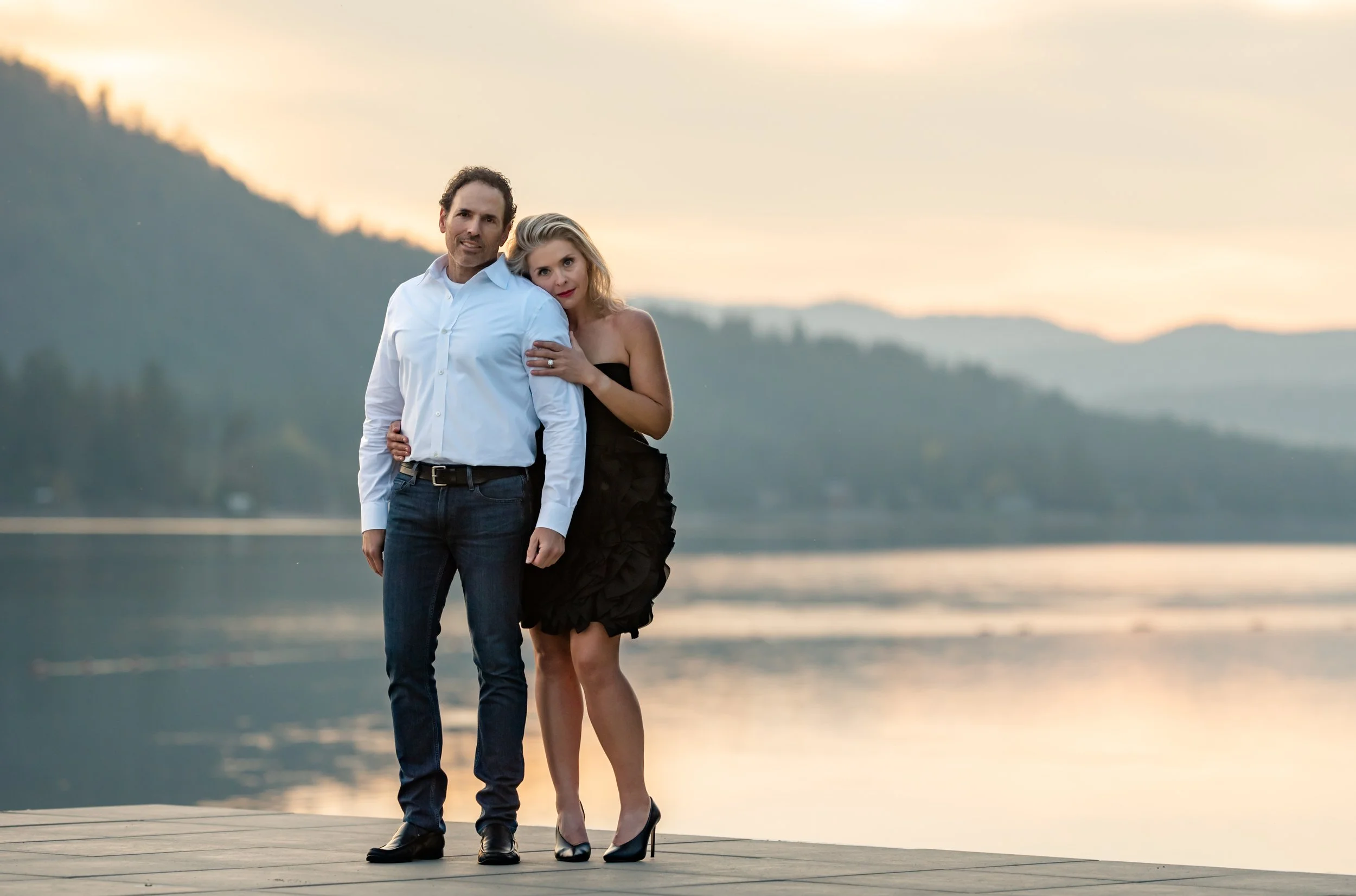 A man and woman stand close together on a dock at sunset, with the Pend Oreille River and hills in the background. The man wears a white shirt and jeans, and the woman wears a black dress and heels, with her arms around his shoulder.