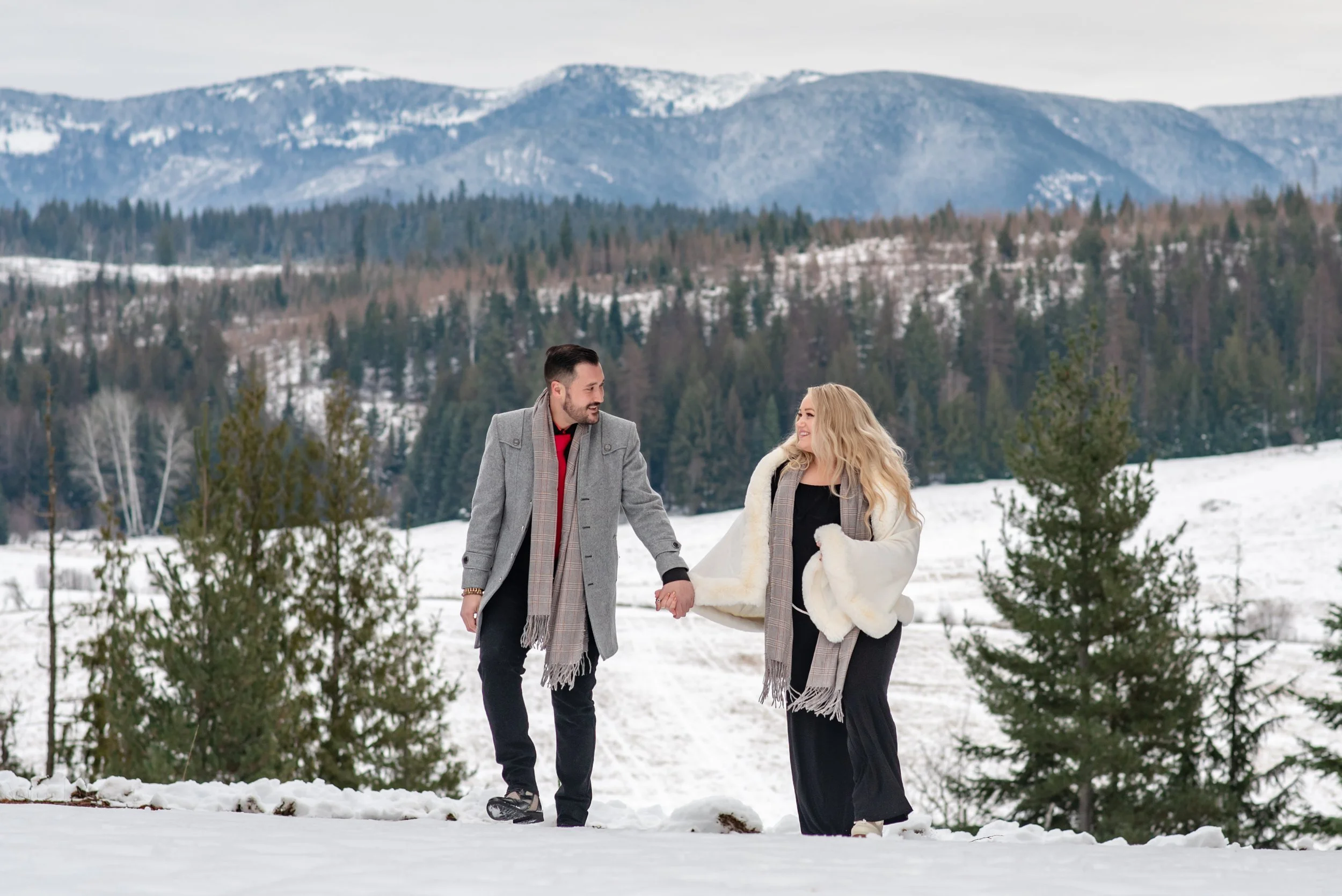 A man and woman holding hands and walking in a snowy landscape with evergreen trees and mountains in the background at Western Pleasure Guest Ranch in Sandpoint, Idaho.