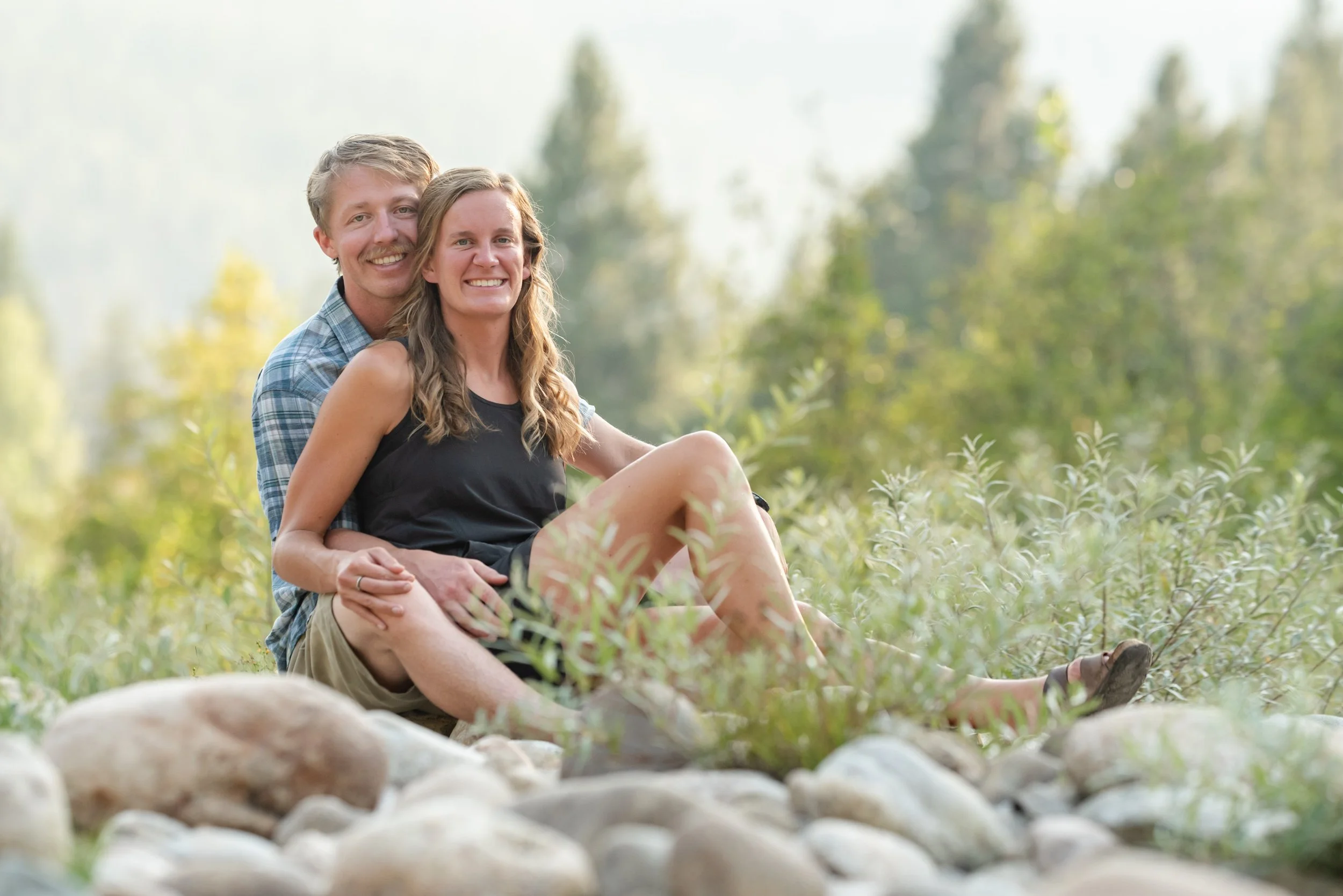 A couple sitting on a rocky surface in an outdoor setting surrounded by greenery, smiling at the camera.