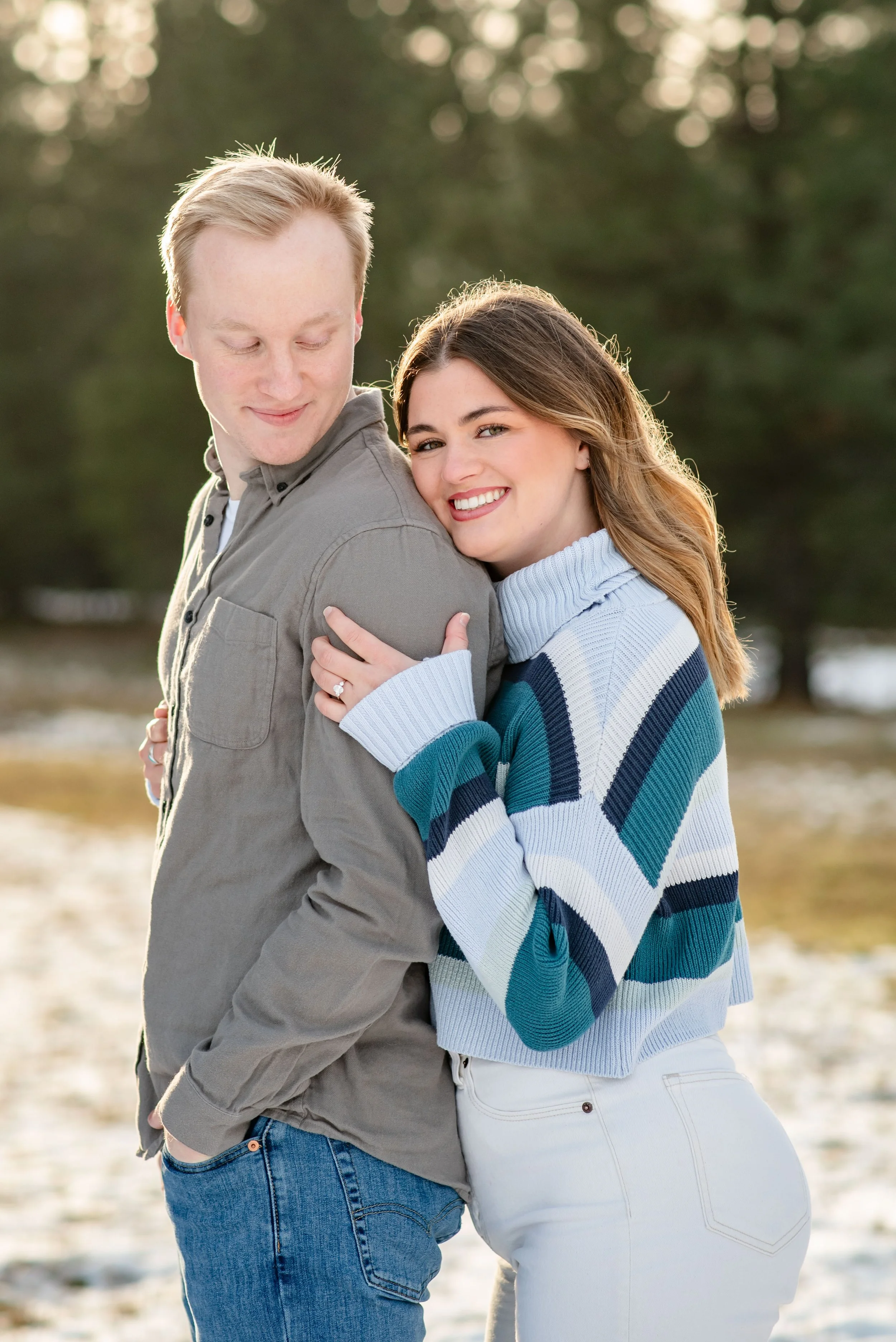 A young man and woman smiling outdoors, with the woman hugging the man from behind in a snowy landscape with trees.