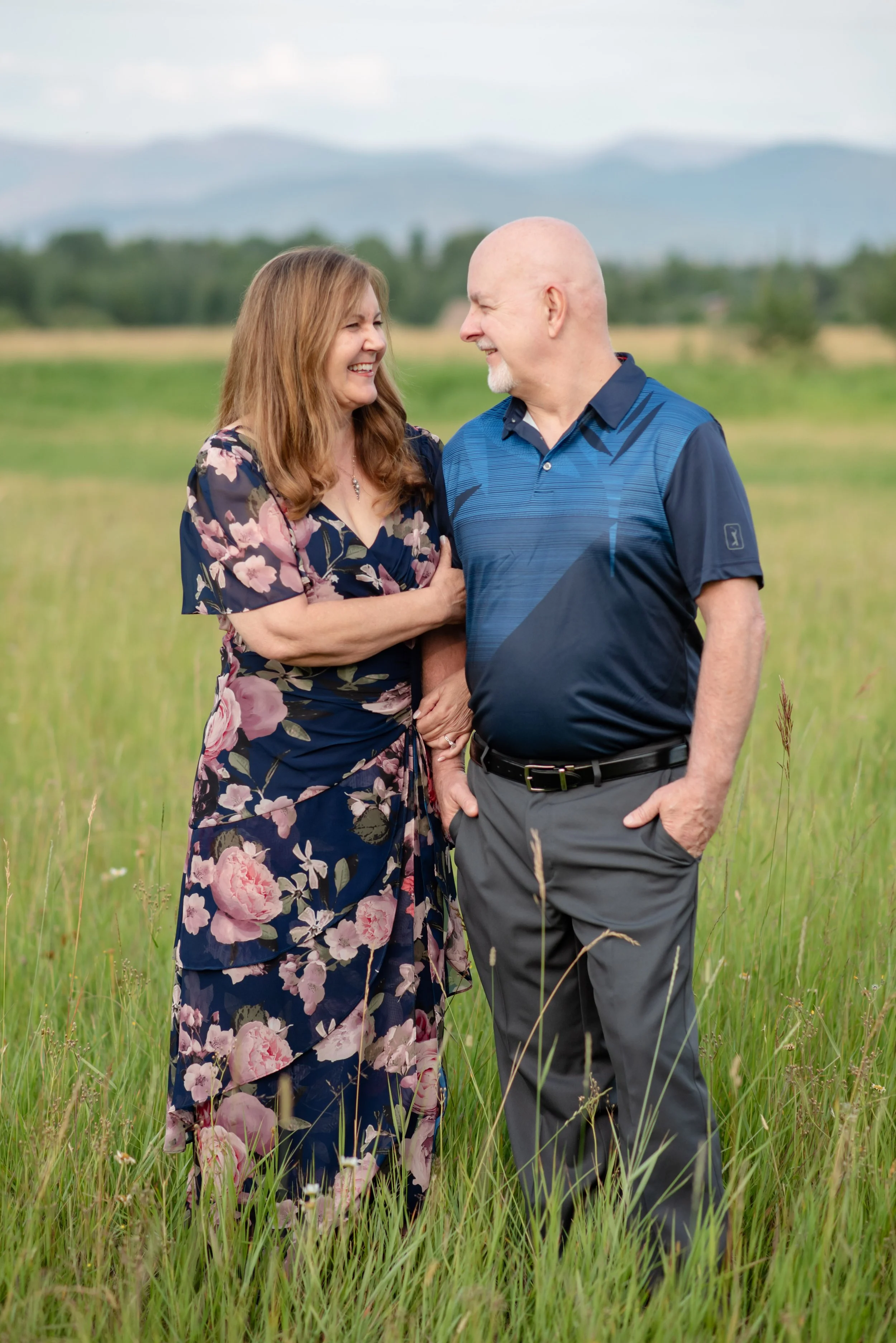 A happy middle-aged couple standing in a field, smiling and looking at each other, with green grass and mountains in the background.
