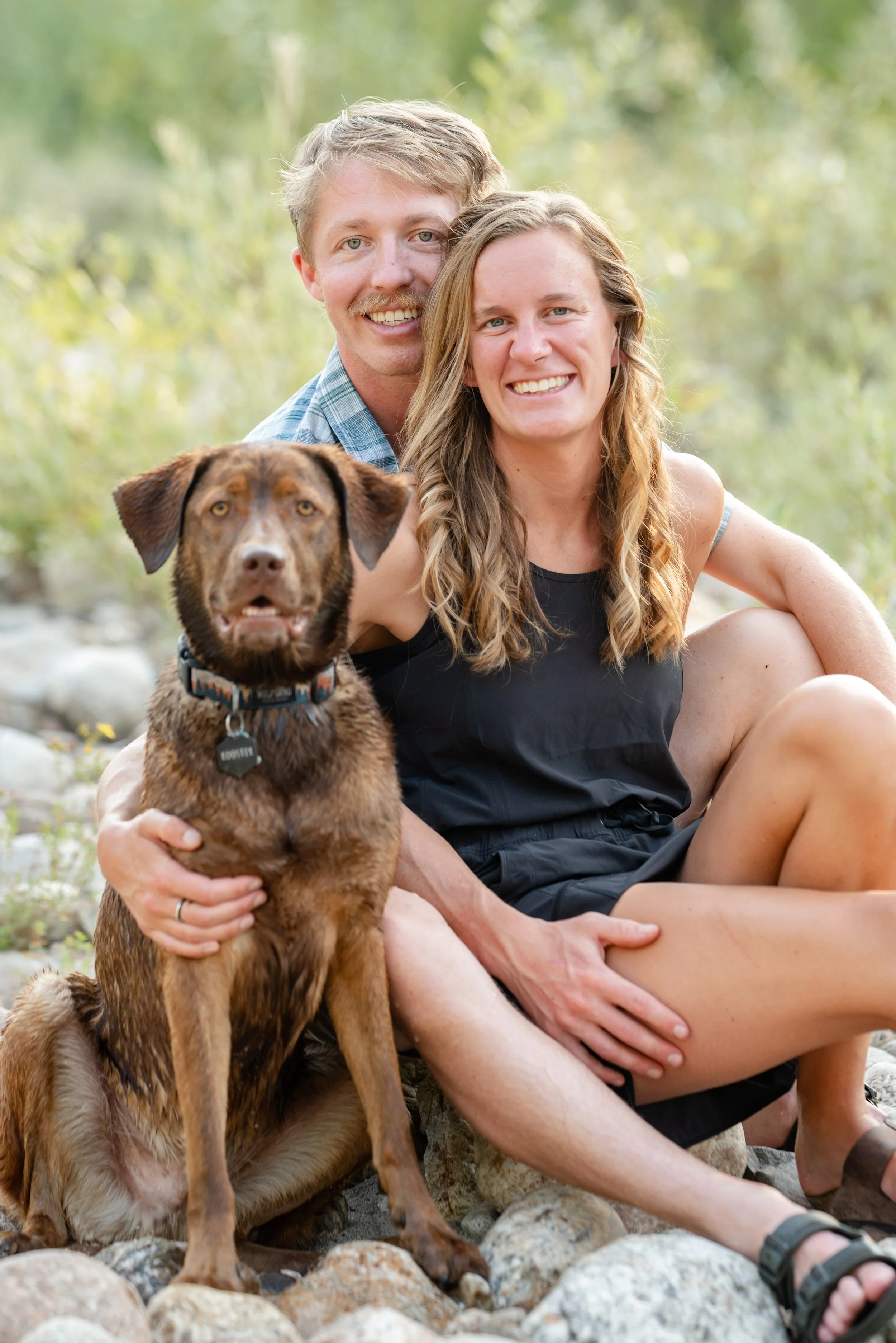 A smiling couple sitting on rocks outdoors with a brown dog in front of them. The man is behind and the woman is sitting to the side, both with relaxed expressions, surrounded by greenery.