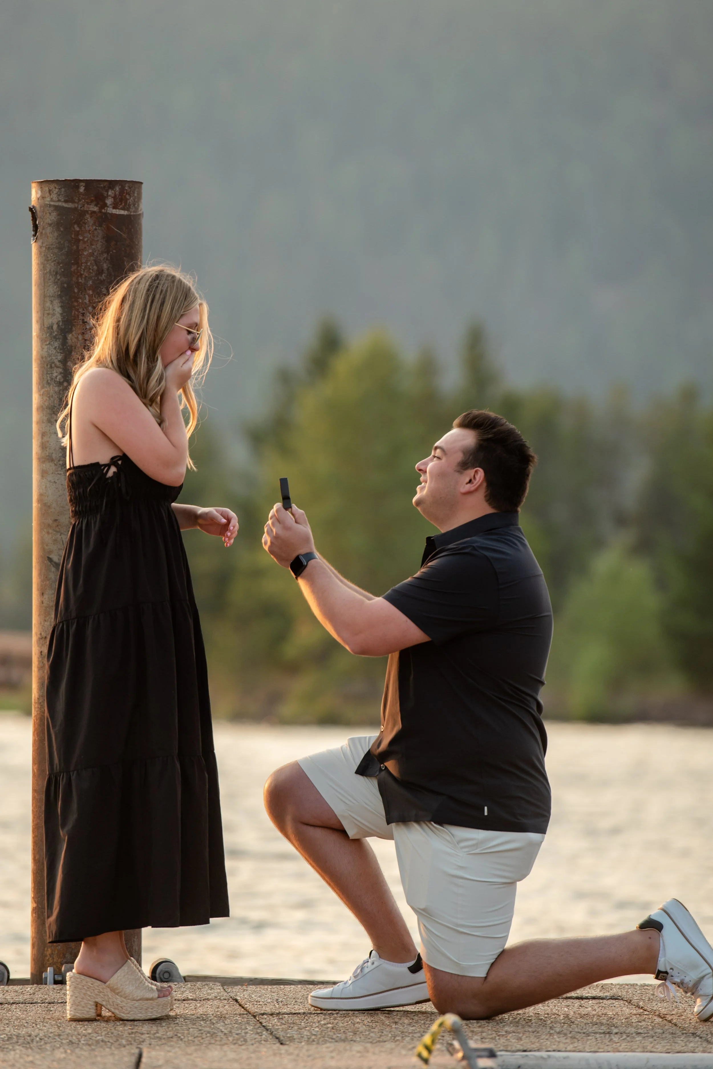 A man proposing marriage to a woman by kneeling on one knee on a dock by the water, while the woman looks surprised and covers her mouth with her hands.