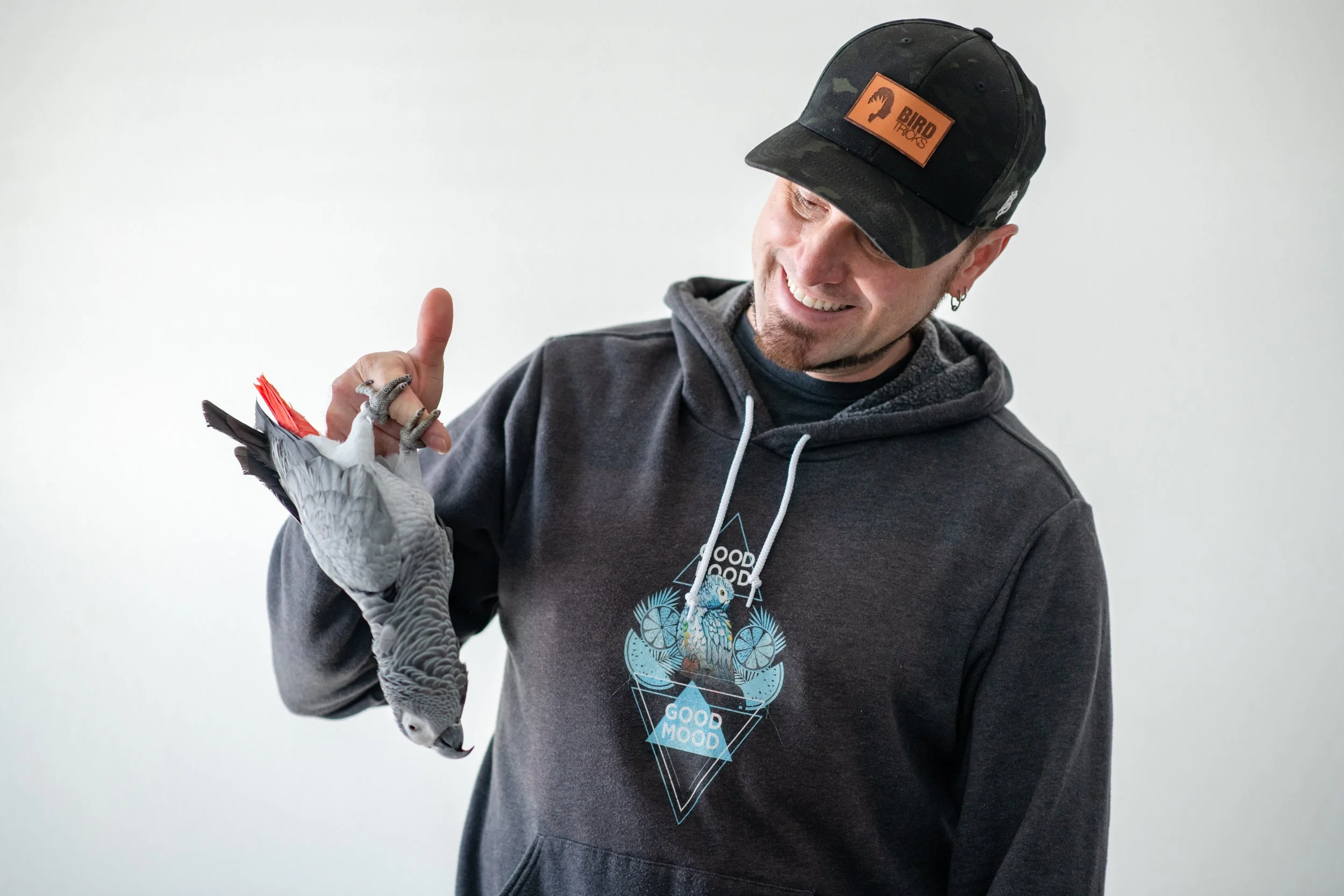 A smiling man in a dark hoodie and baseball cap holding a small African Grey bird.
