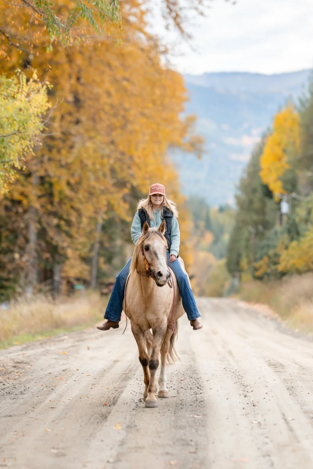 A woman riding a light-colored horse on a dirt road surrounded by autumn trees with yellow and orange leaves.