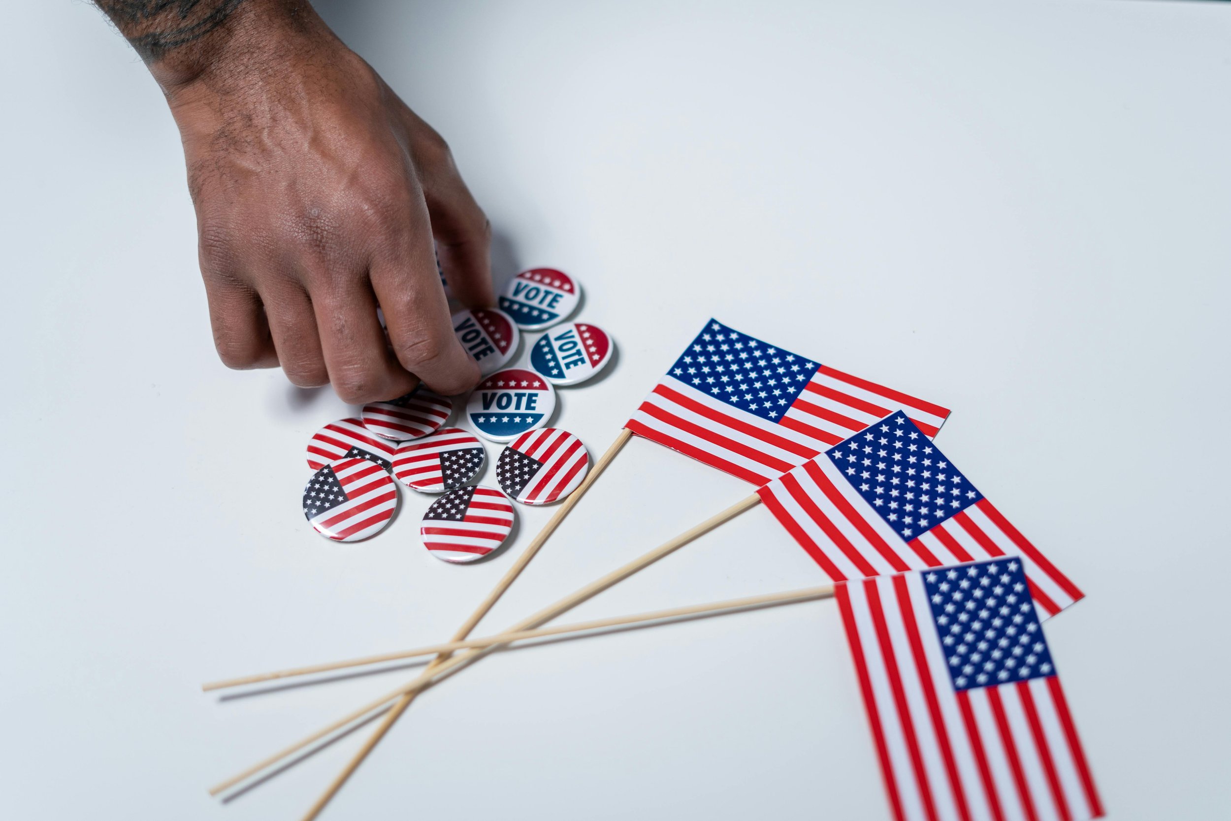 A hand reaches for a pile of red, white, and blue “VOTE” buttons on a table beside several small American flags. Reflecting the perspective of Kevin Wall on campaign volunteering.