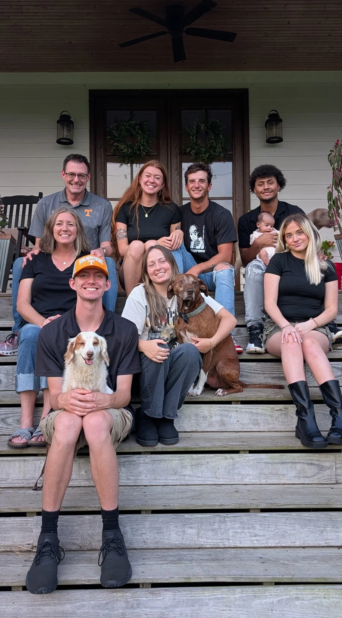A group photo of ten people and two dogs on the steps of a house, with some people smiling and others with neutral expressions. The people appear to be of various ages and backgrounds, and the house has a porch with coat hooks and wreaths on the door.