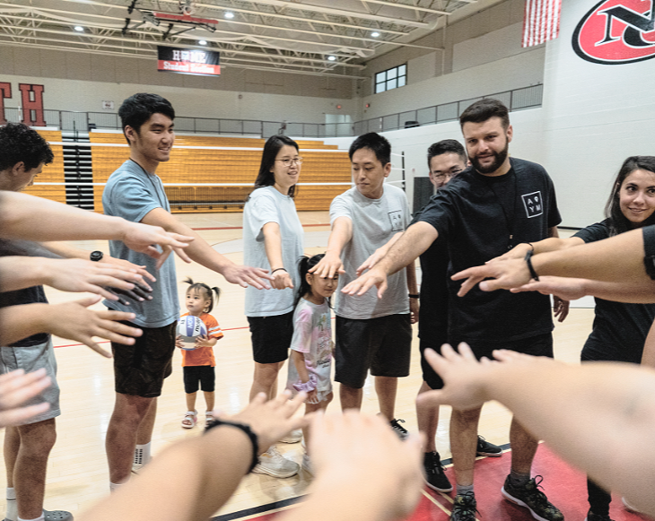 A group of people, including children, standing in a circle in a gymnasium, reaching towards the center in a team-building activity.