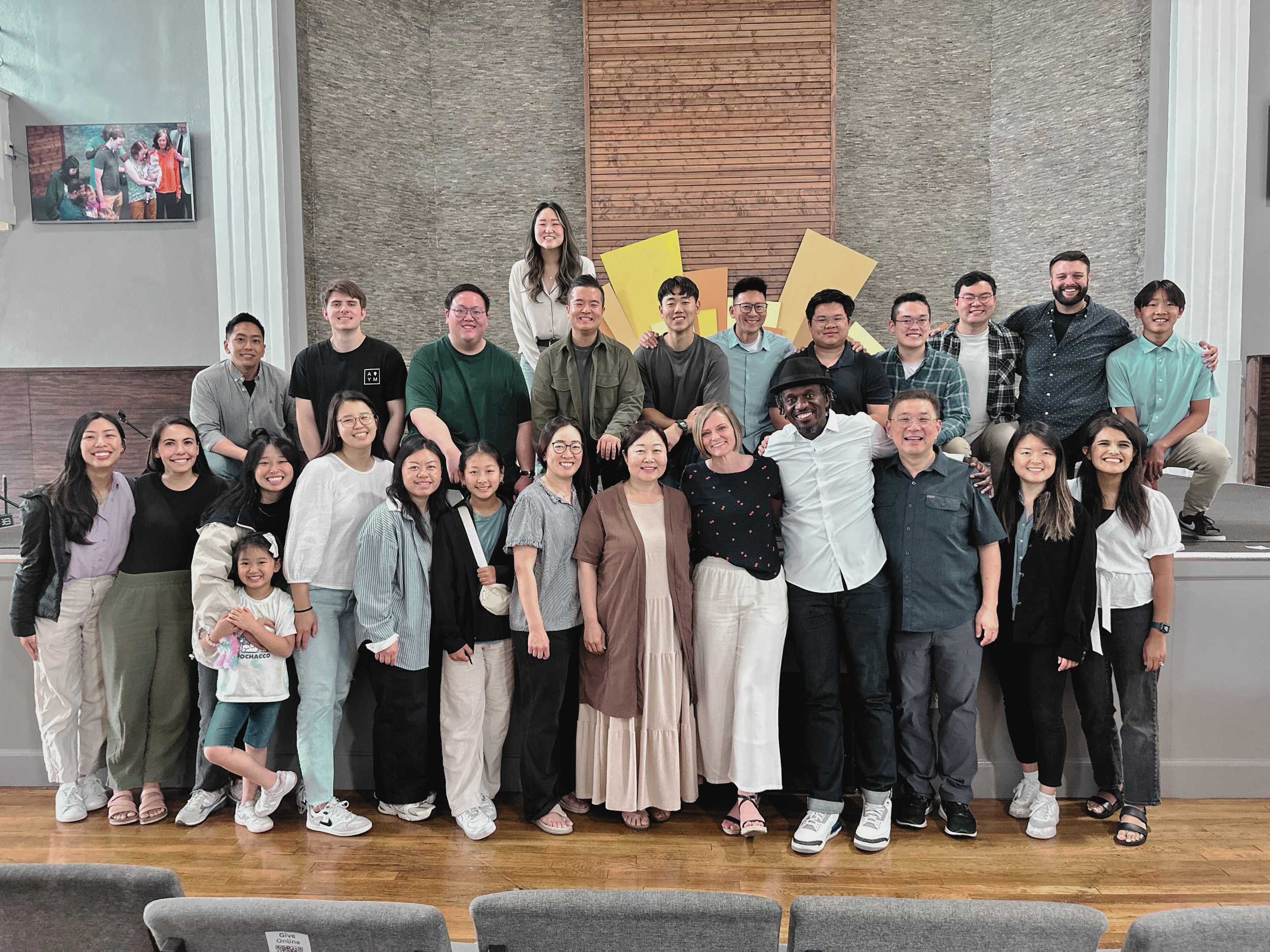 Group photo of diverse people in a room with wooden floors, two rows of standing and sitting, smiling. Background has a textured wall and artwork. A screen on the left displays a smaller image.