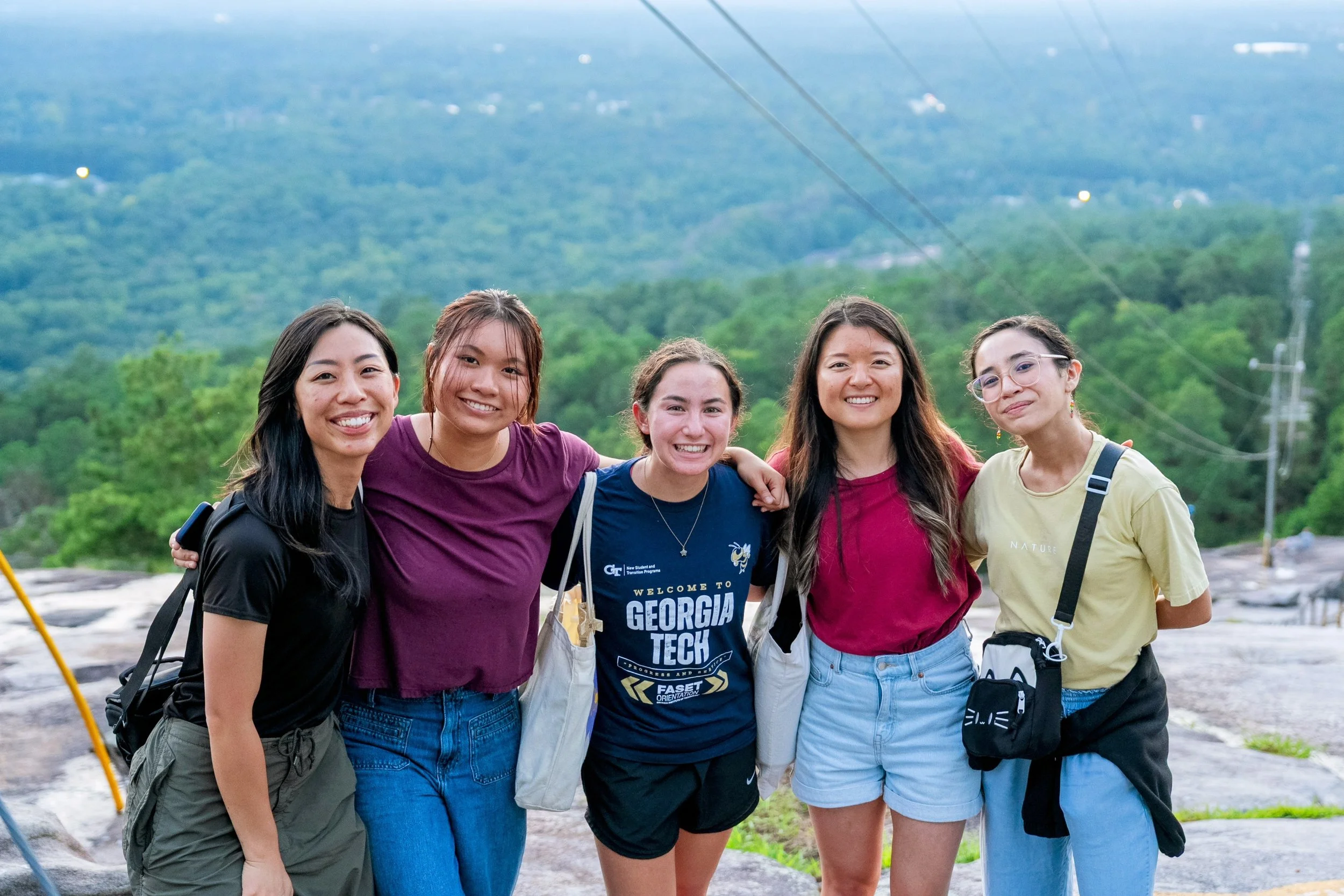 Five young women smiling with arms around each other outdoors in front of lush green forest, mountains in the background, and power lines overhead.