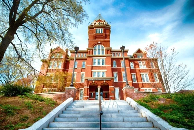 A historic building with a tower at Georgia Tech University, set against a partly cloudy sky, with white stone stairs leading up to its entrance and trees surrounding it.