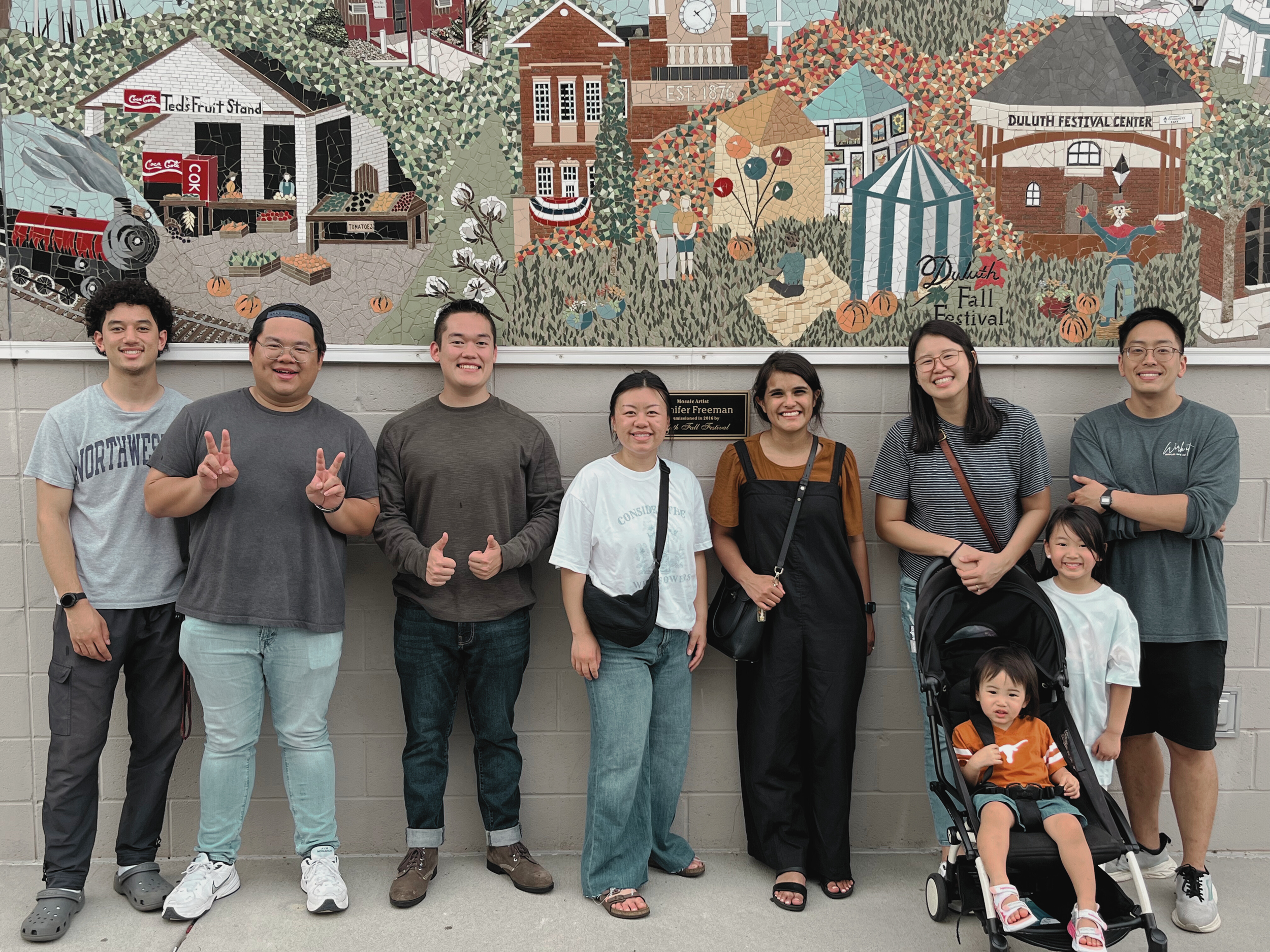 Group of nine people, including children, standing in front of a mural with a watercolor scene depicting a fall festival, a train, and buildings in downtown Atlanta
