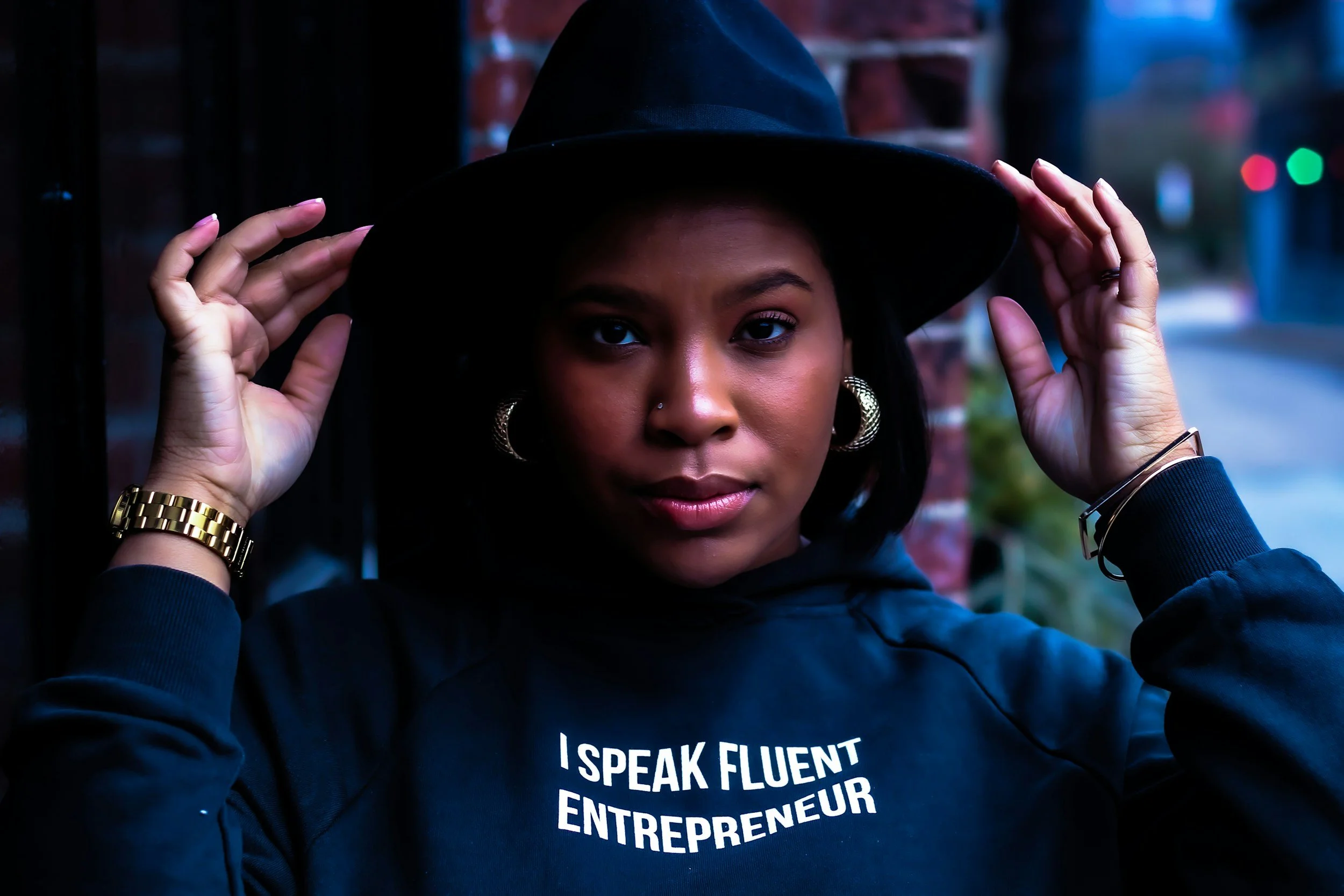 A woman wearing a black hat and hoodie with the text "I SPEAK FLUENT ENTREPRENEUR" embroidered on it, standing outdoors near a brick wall, with a city street in the background.