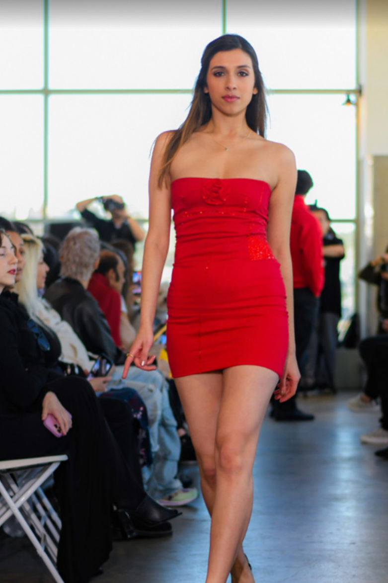 A woman in a red strapless dress walking on a runway during a fashion show, with an audience seated on both sides.