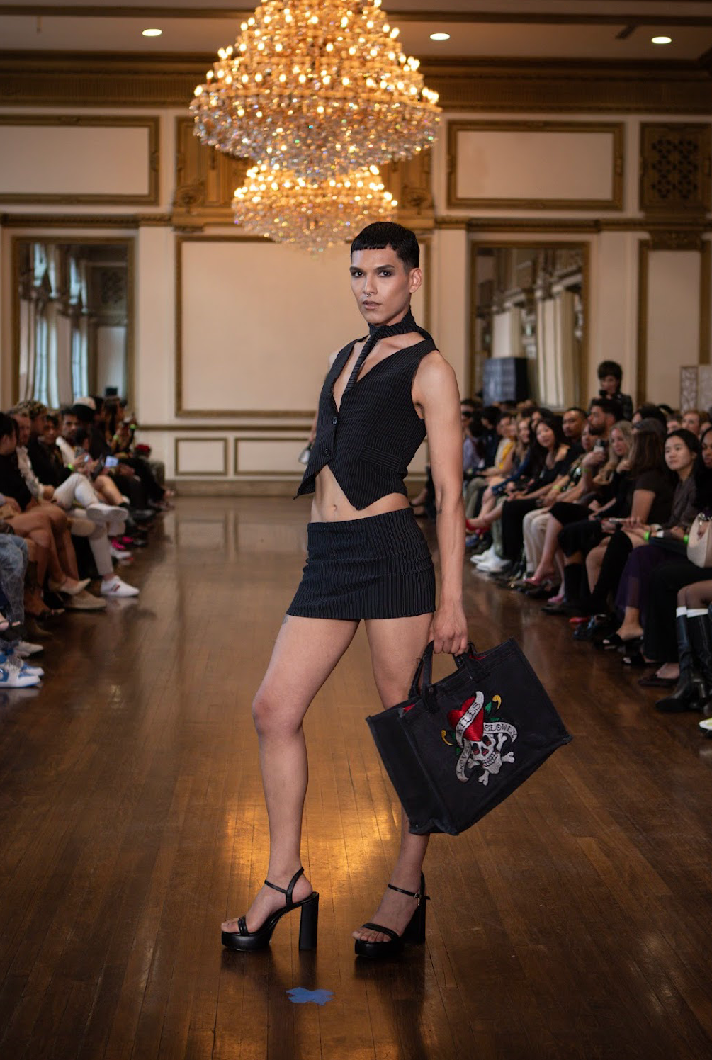 A female model walking in a fashion show, wearing a black sleeveless vest and matching high-waisted mini skirt, high heels, and carrying a black bag with a skull and snake design. The show is indoors with an audience watching and a large chandelier o