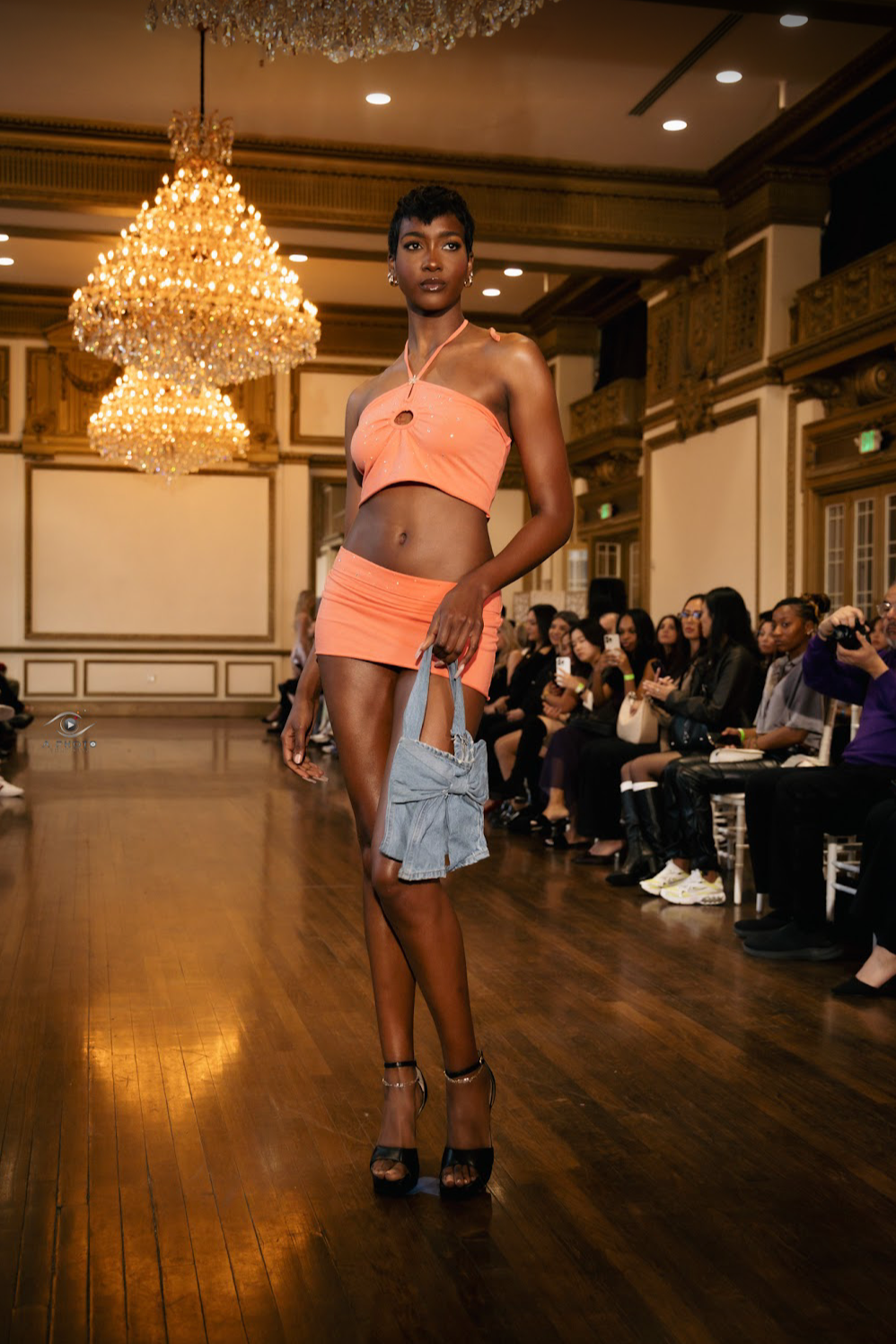 A female model walks down a runway in a peach two-piece outfit, carrying a denim bag, with chandeliers hanging above and an audience sitting on either side taking photos.