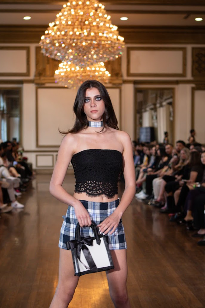 A young woman walking on a runway at a fashion show, wearing a strapless black lace top, plaid shorts, and holding a small black and white handbag with a ribbon. The runway is in a grand room with a large chandelier above and an audience seated on bo