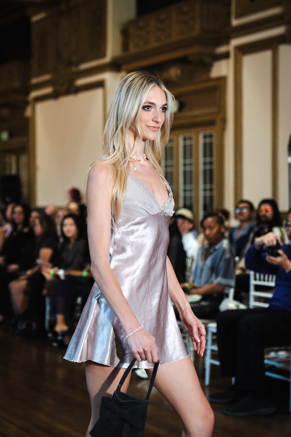 A young woman walking on a runway at a fashion show, wearing a satin slip dress and holding a small black purse. She has long blonde hair and is wearing jewelry, with an audience seated in the background.
