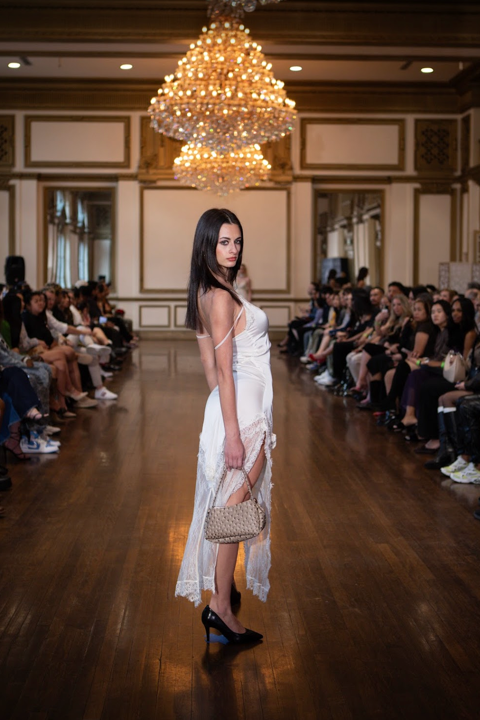 A female model walking down the runway at a fashion show, wearing a white slip dress with lace details, black high heels, holding a beige handbag, with an audience seated on both sides, and a chandelier hanging overhead in an elegant room.