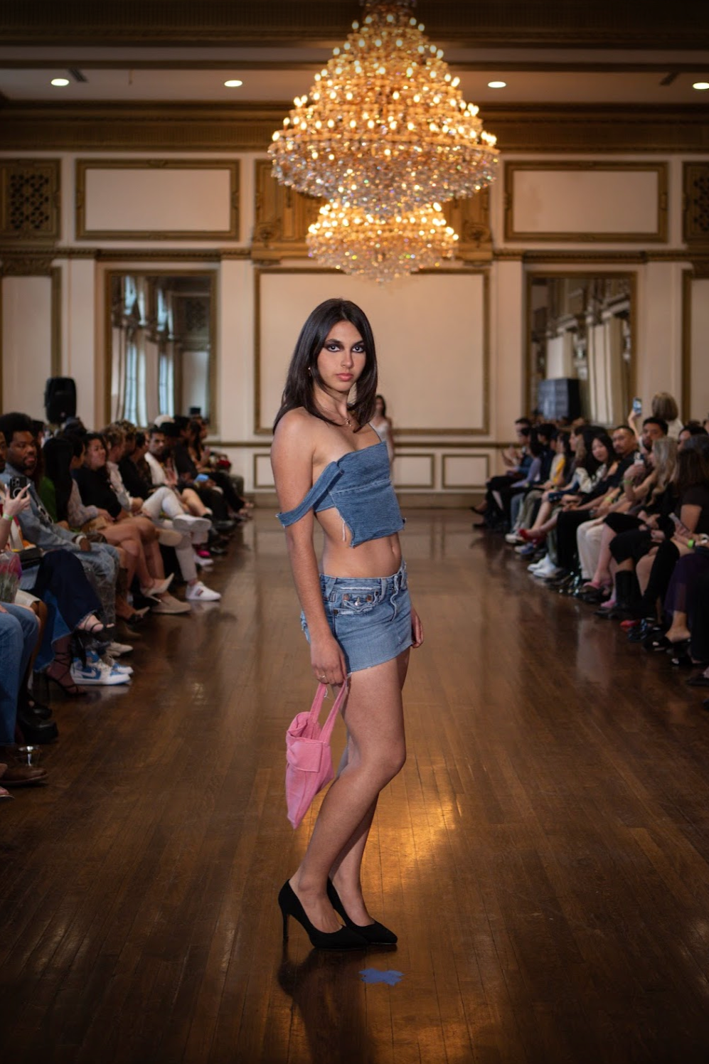 A young woman walking down the runway at a fashion show, wearing a casual denim crop top and skirt, holding a pink bag, with an elegant chandelier hanging above and audience seated along both sides of the runway.