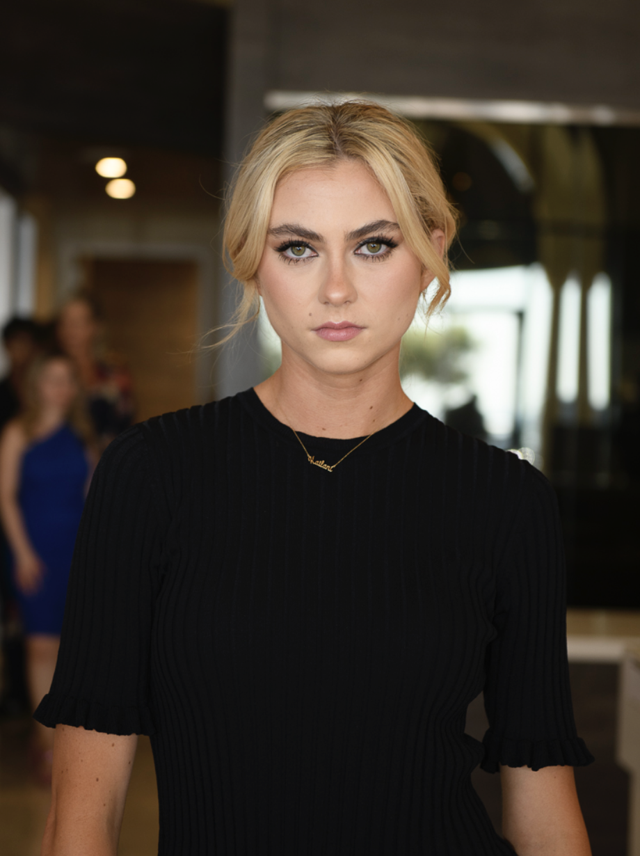 A young woman with blonde hair pulled back, wearing a black ribbed top with short ruffled sleeves, looking directly at the camera in an indoor setting with people in the background.