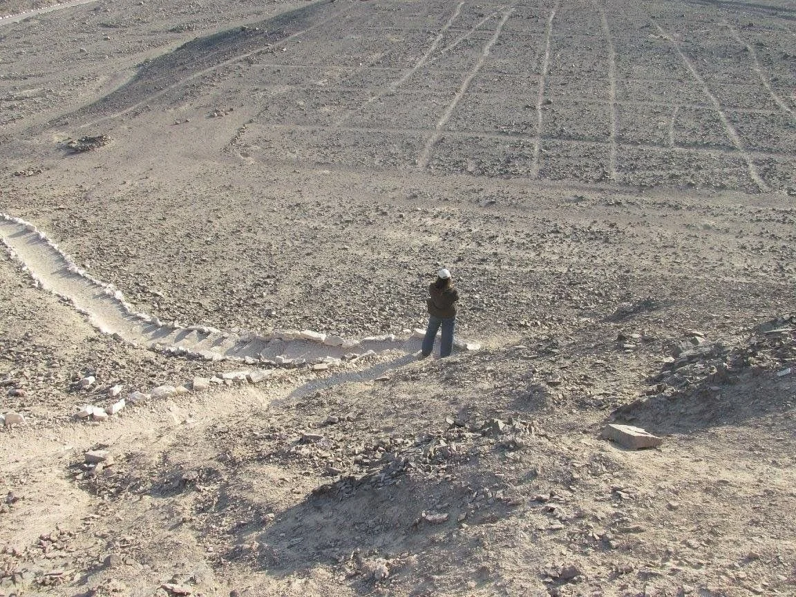 A person standing on a rugged dirt terrain with a rock-lined path or boundary in an arid, rocky landscape.