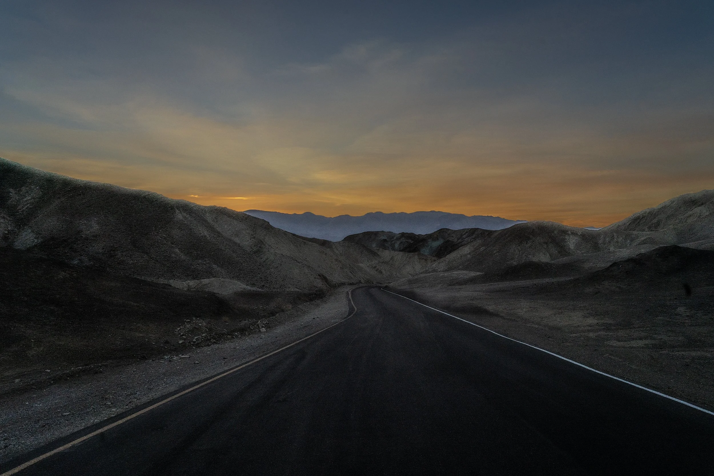 Open road winding through mountainous desert landscape at sunset, with a colorful sky and distant mountain ranges.