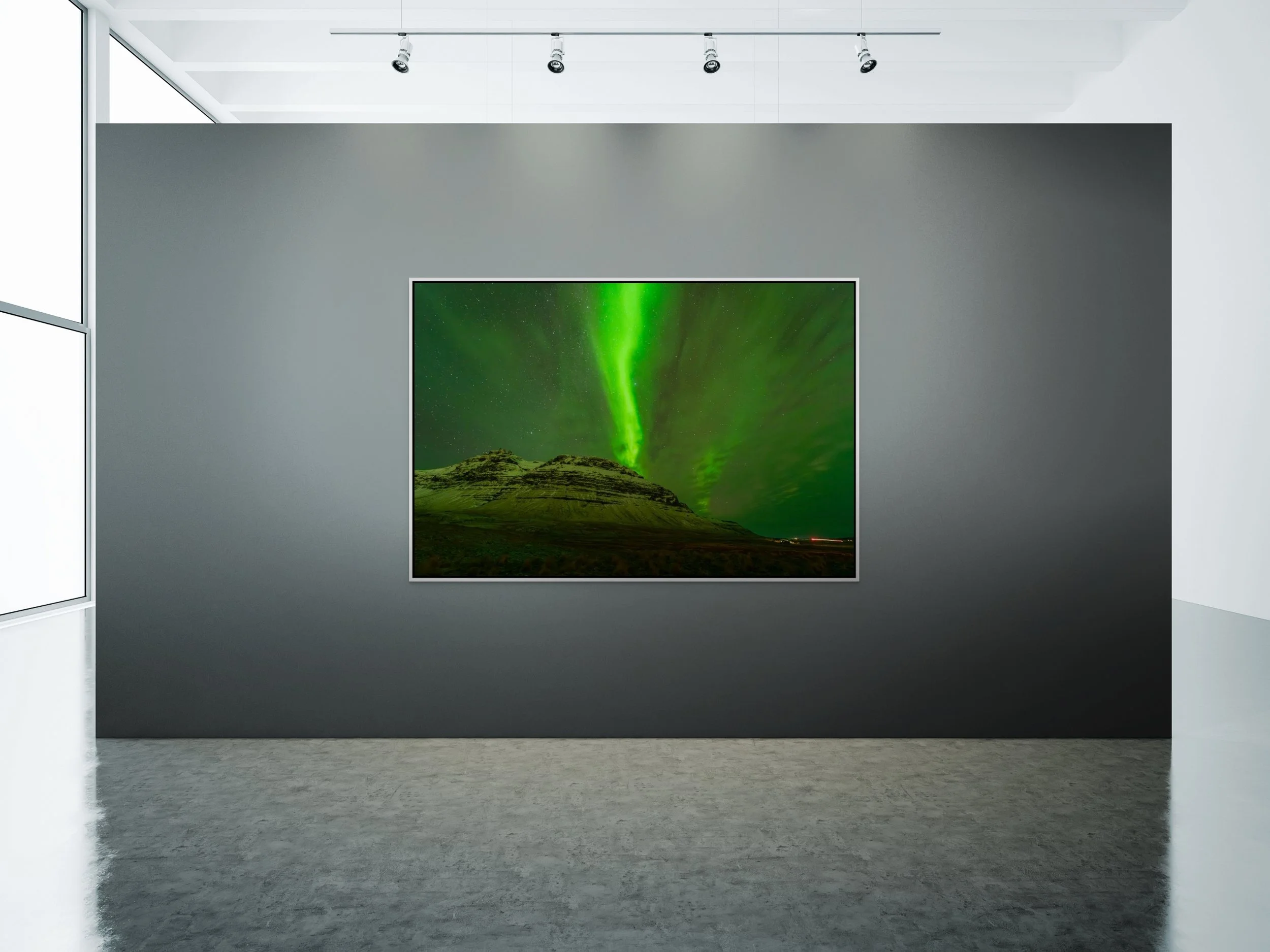 Veil of Light — green aurora over an Iceland peak, framed in white floating wood frame on a wide gallery wall with ceiling track lighting and polished concrete floor.