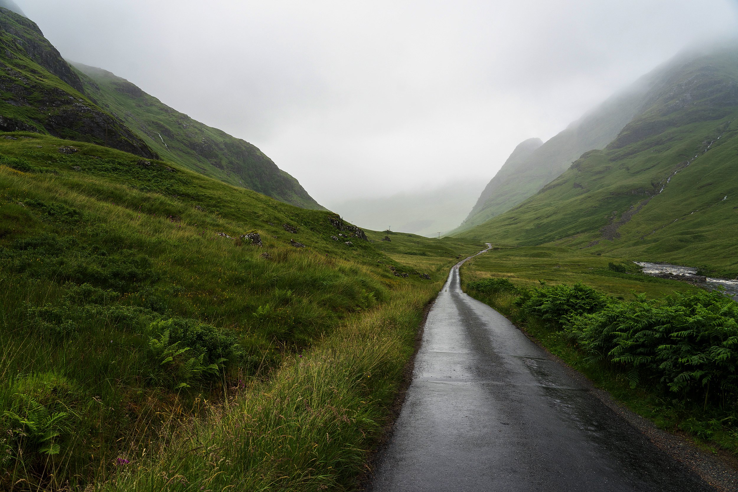 Rain-slick single-track road vanishing into fog between green slopes in Glen Etive, Scottish Highlands. Skyfall James Bond 007 filming location.