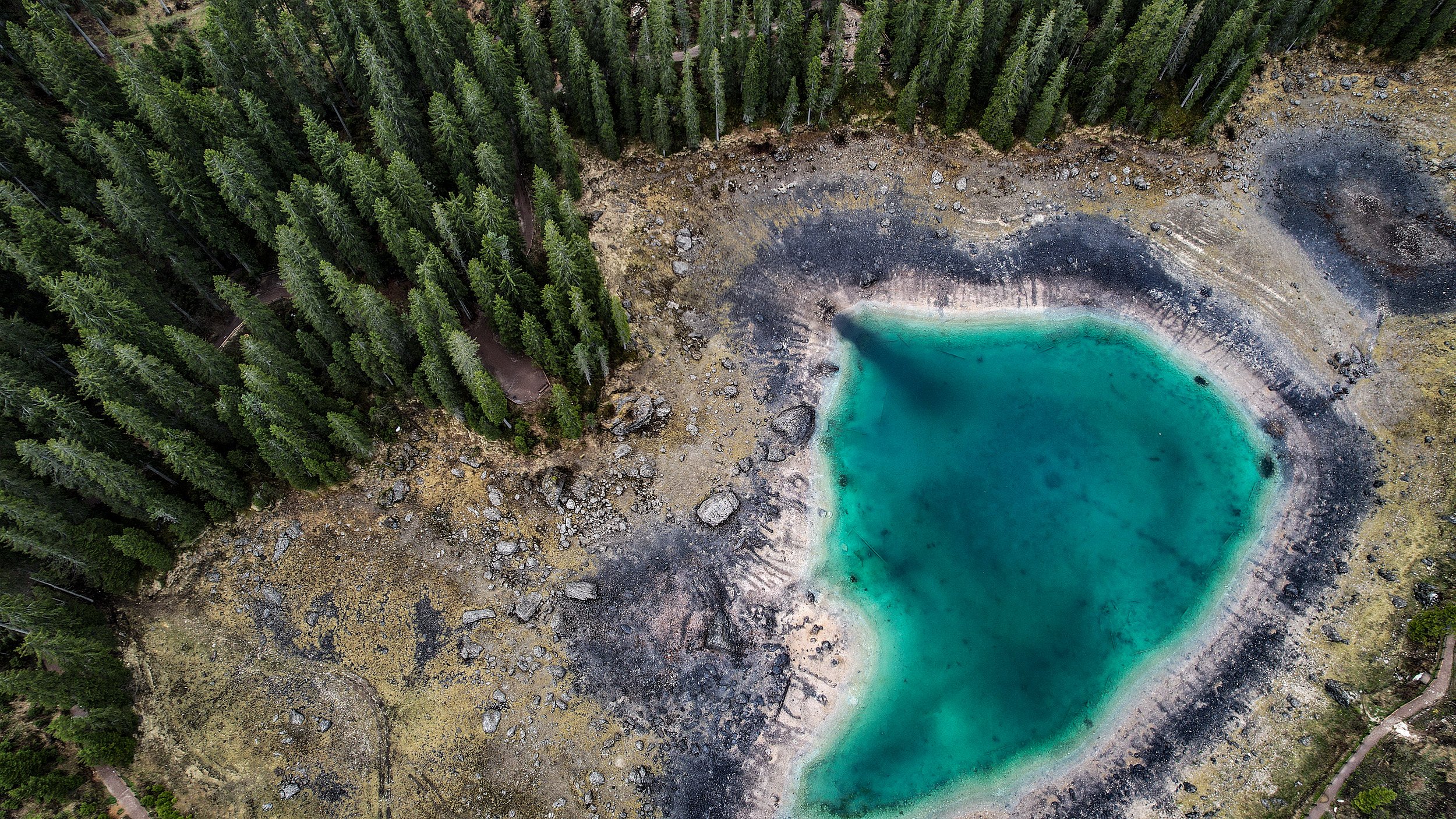 An aerial view of a small, turquoise pond surrounded by a rocky and forested landscape, with dense green trees on one side.
