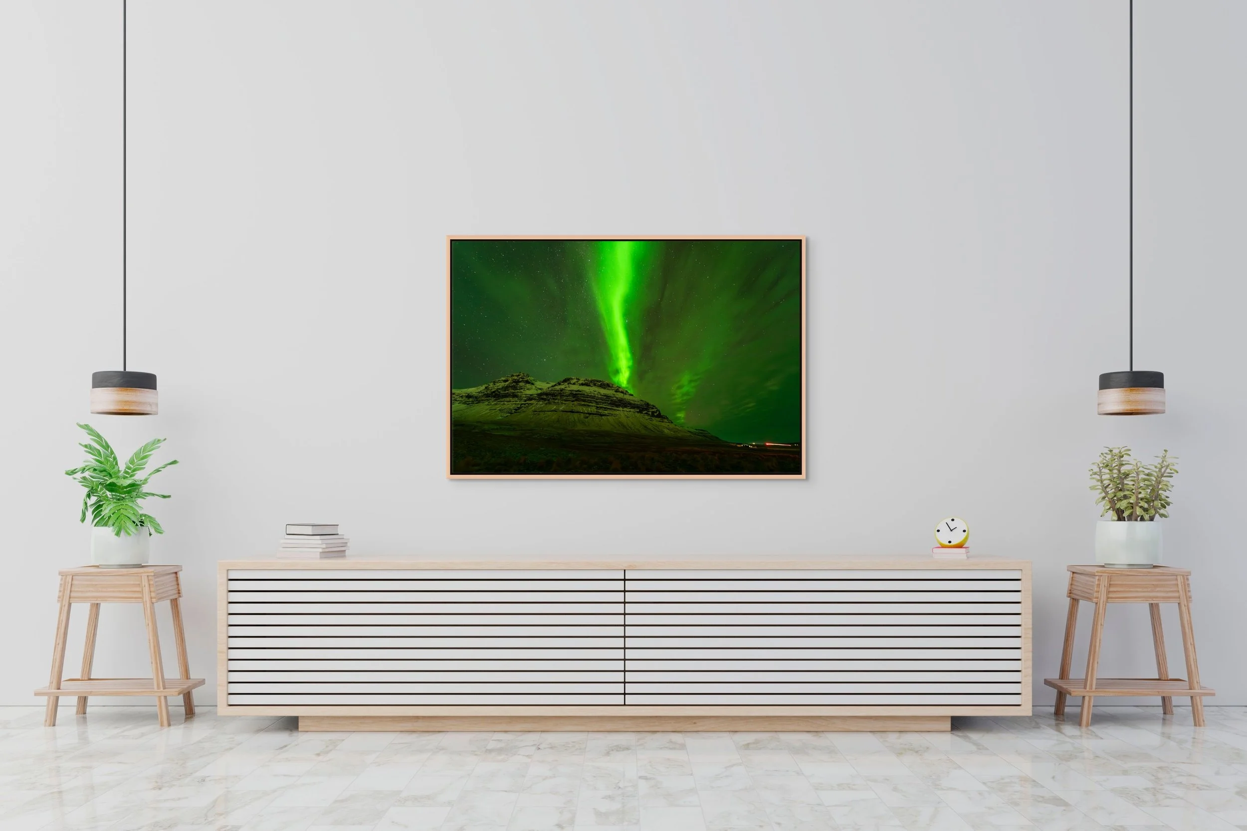 Veil of Light — green aurora over an Iceland peak, framed in oak floating wood frame above a minimalist white credenza with twin sconces and potted plants.