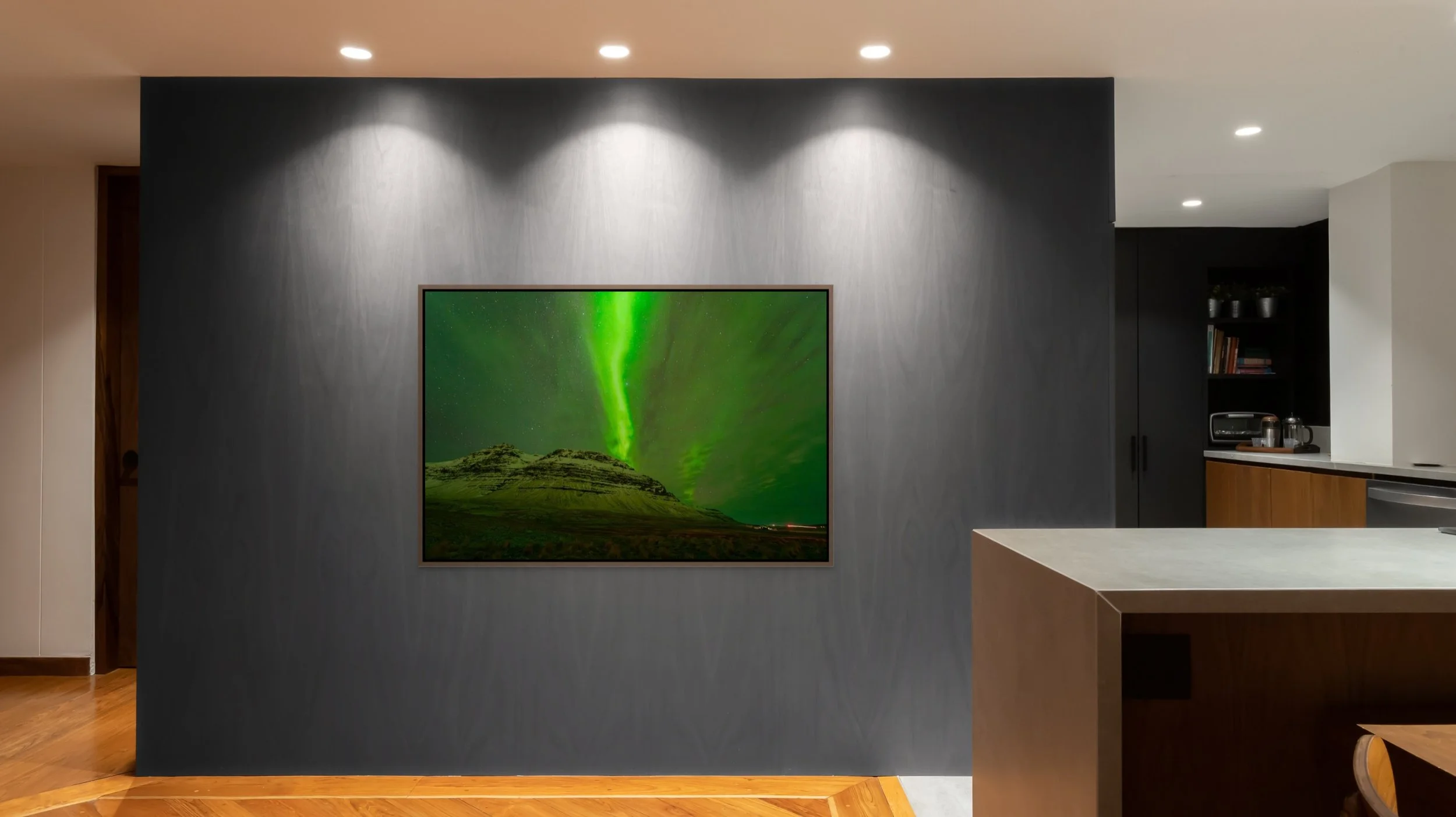 Veil of Light — green aurora over an Iceland peak, framed in alder brown floating wood frame on a charcoal feature wall with recessed spotlights beside a modern kitchen.