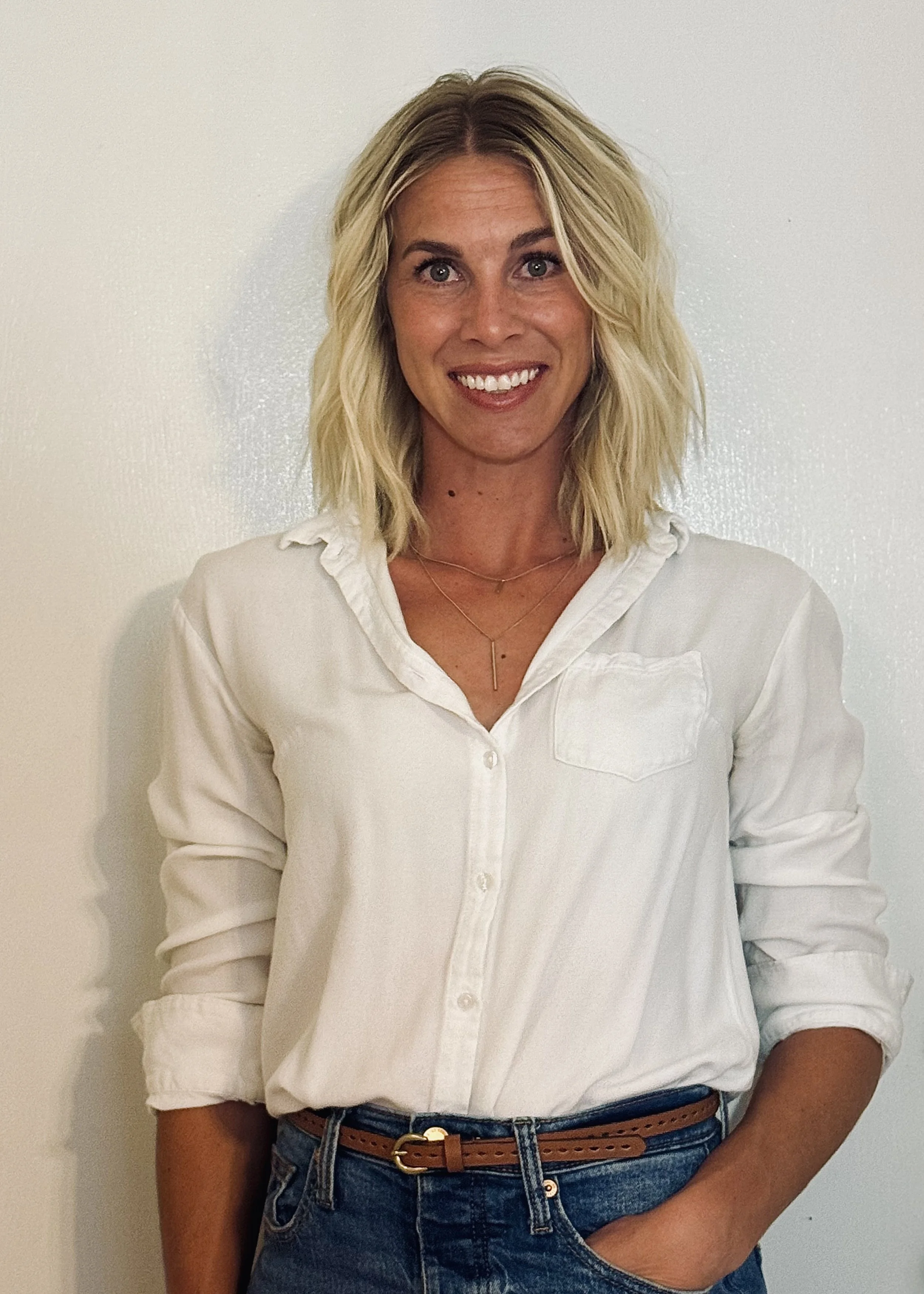 A woman with blonde, shoulder-length wavy hair, wearing a white button-up shirt with rolled-up sleeves, blue jeans with a brown belt, smiling and standing against a plain white wall.