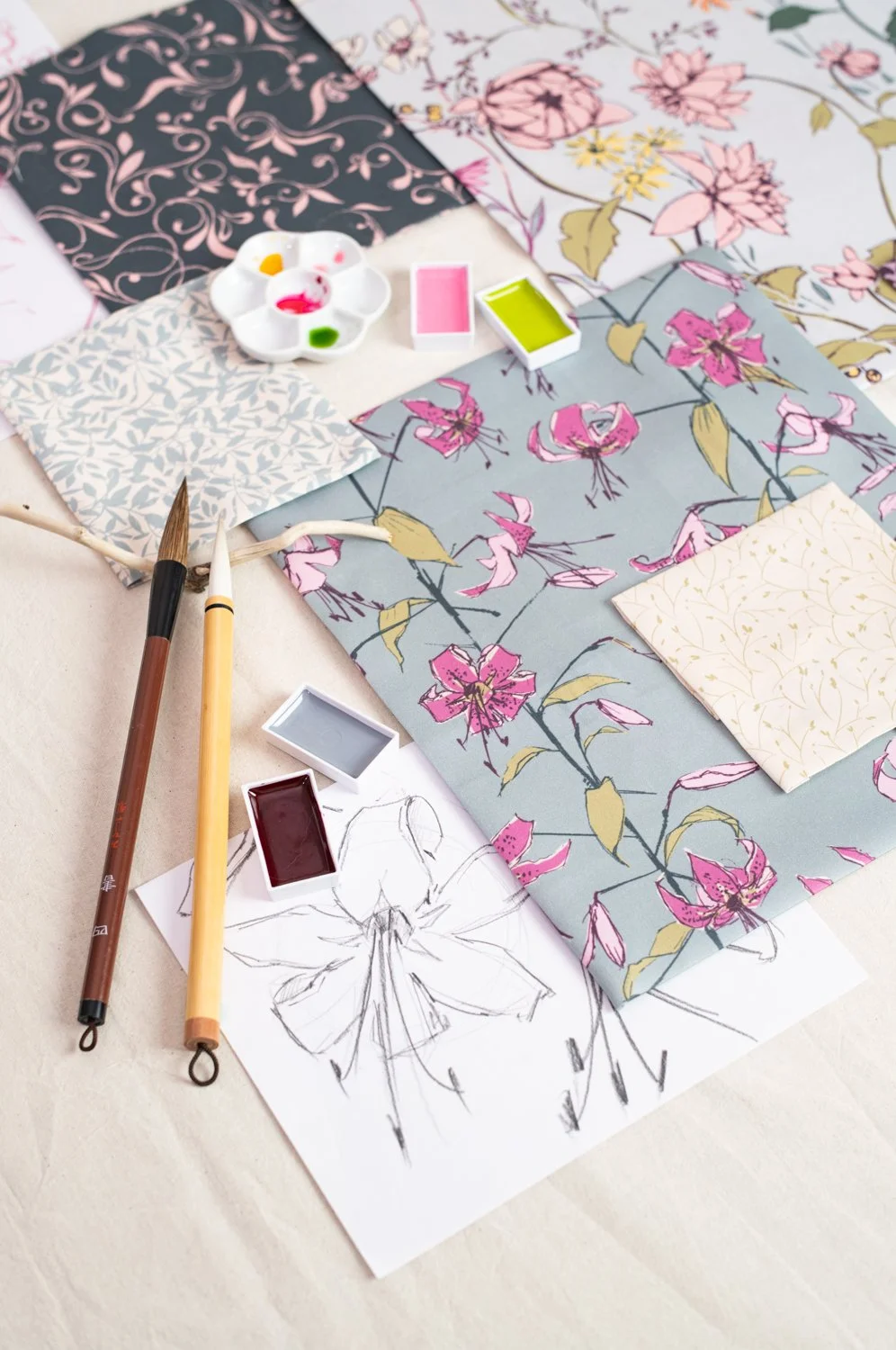 Artistic workspace with floral patterned papers, watercolor paints, brushes, and a sketch of a flower.