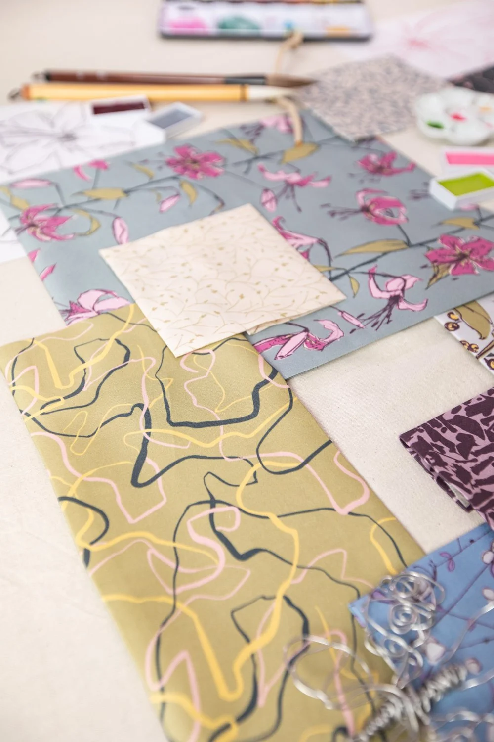 Assorted fabric swatches with floral and abstract patterns, along with sewing tools and color paints on a table.